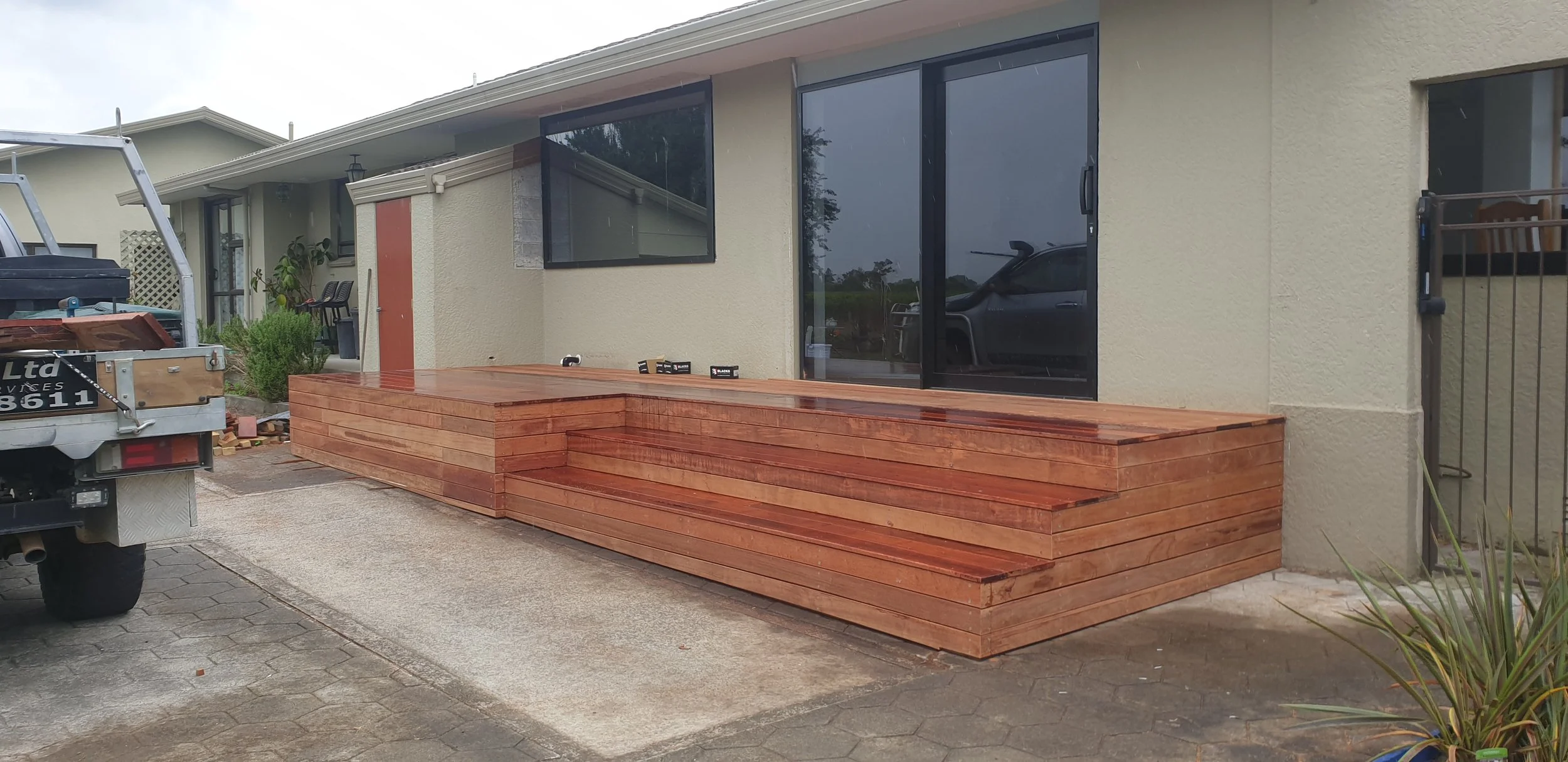 A newly built wooden deck attached to the back of a house, with stairs leading down to the driveway. A truck with tools and materials is parked nearby, suggesting ongoing construction. The house has large glass sliding doors, and there is patio furni