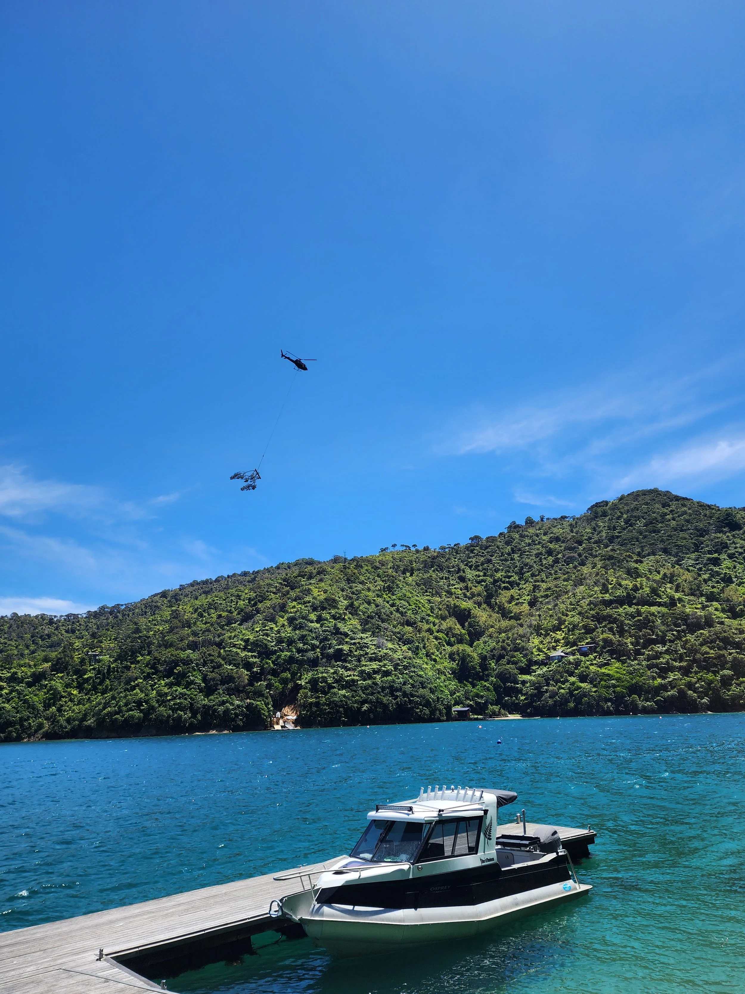 A boat docked at a wooden pier on a body of water, with a green, forested hillside and a bright blue sky in the background. A helicopter is flying in the sky with a cable hanging from it, carrying a load.