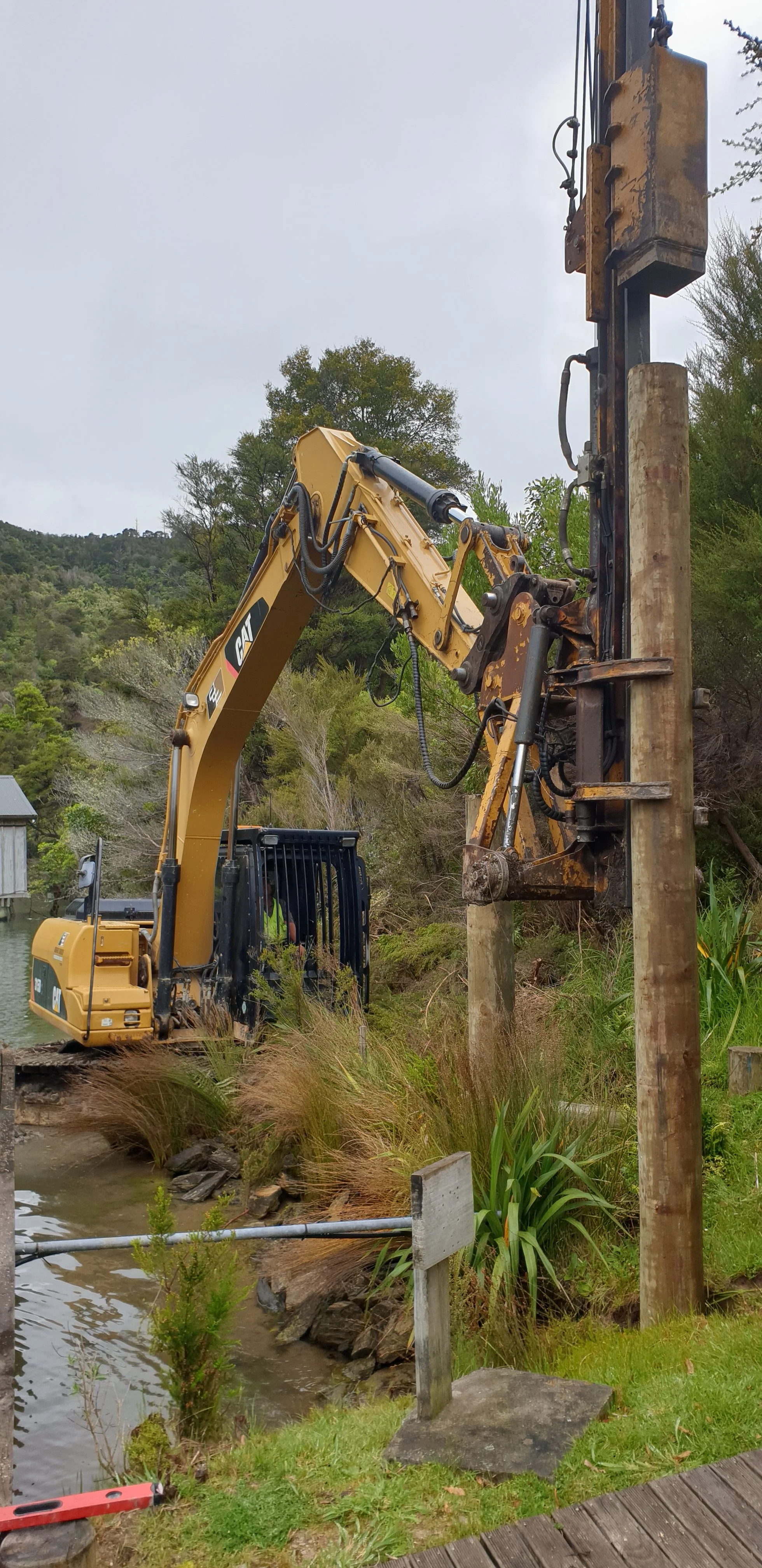A yellow excavator working near a riverbank with a large wooden utility pole, surrounded by greenery and trees.