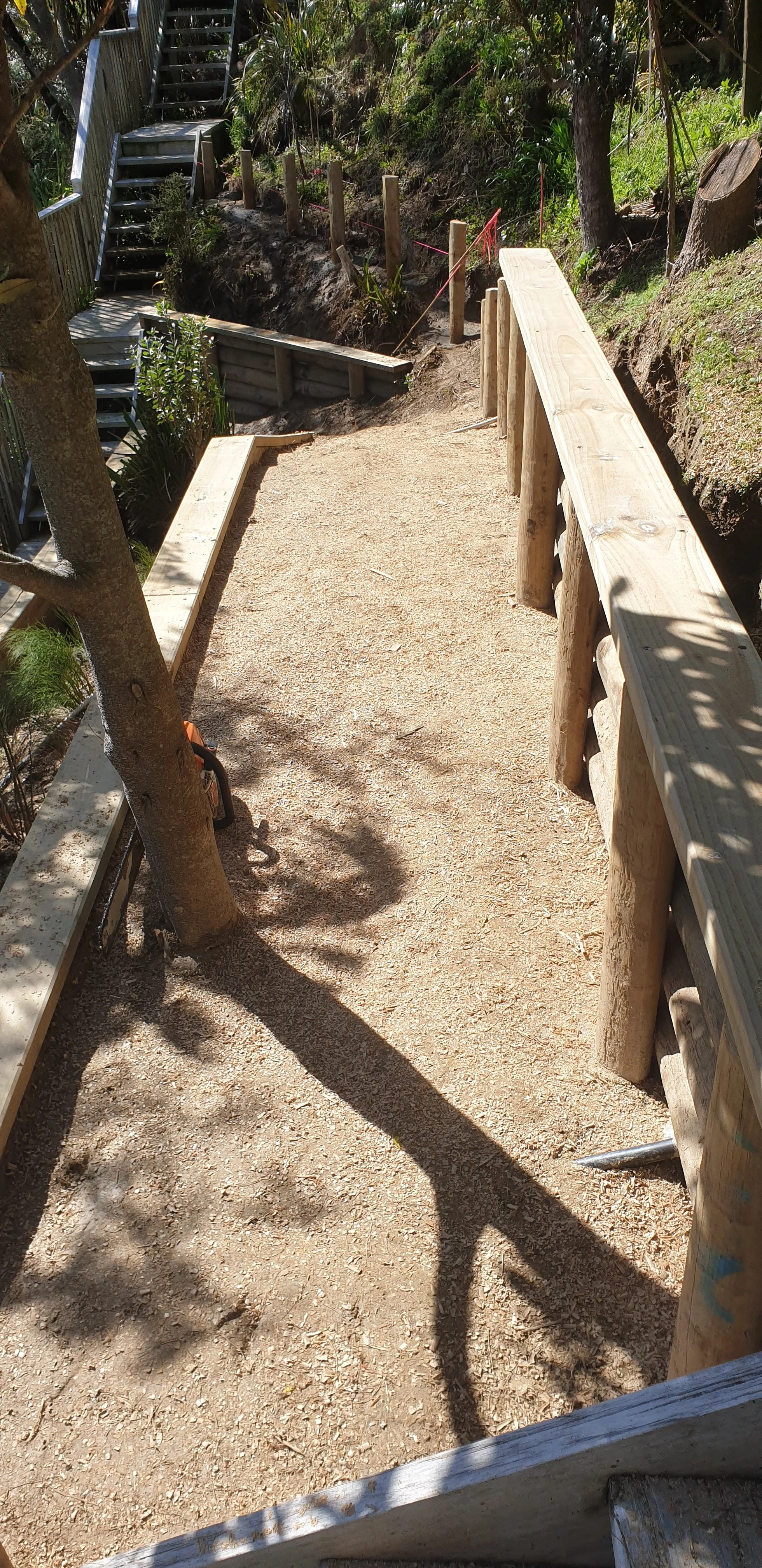 Construction site with a dirt pathway, wooden railings, and a staircase leading up a hillside, surrounded by trees and plants.