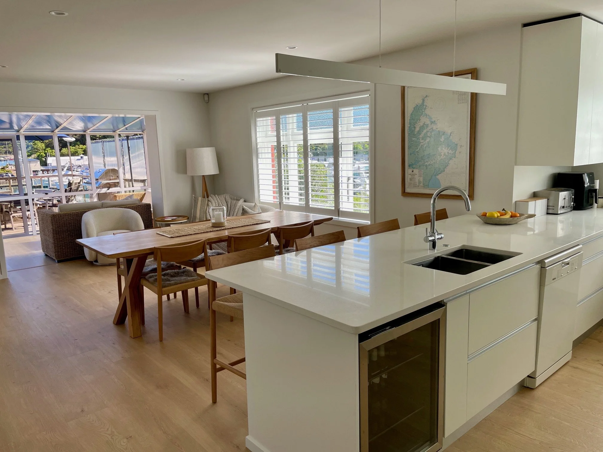 A bright, modern open-concept kitchen and dining area with a view into a living space and a porch outside. The kitchen features a large white island with a built-in sink and a bowl of fruit, and there are wooden chairs around the dining table. The living area has a wicker armchair, a white armchair, and large windows with shutters.
