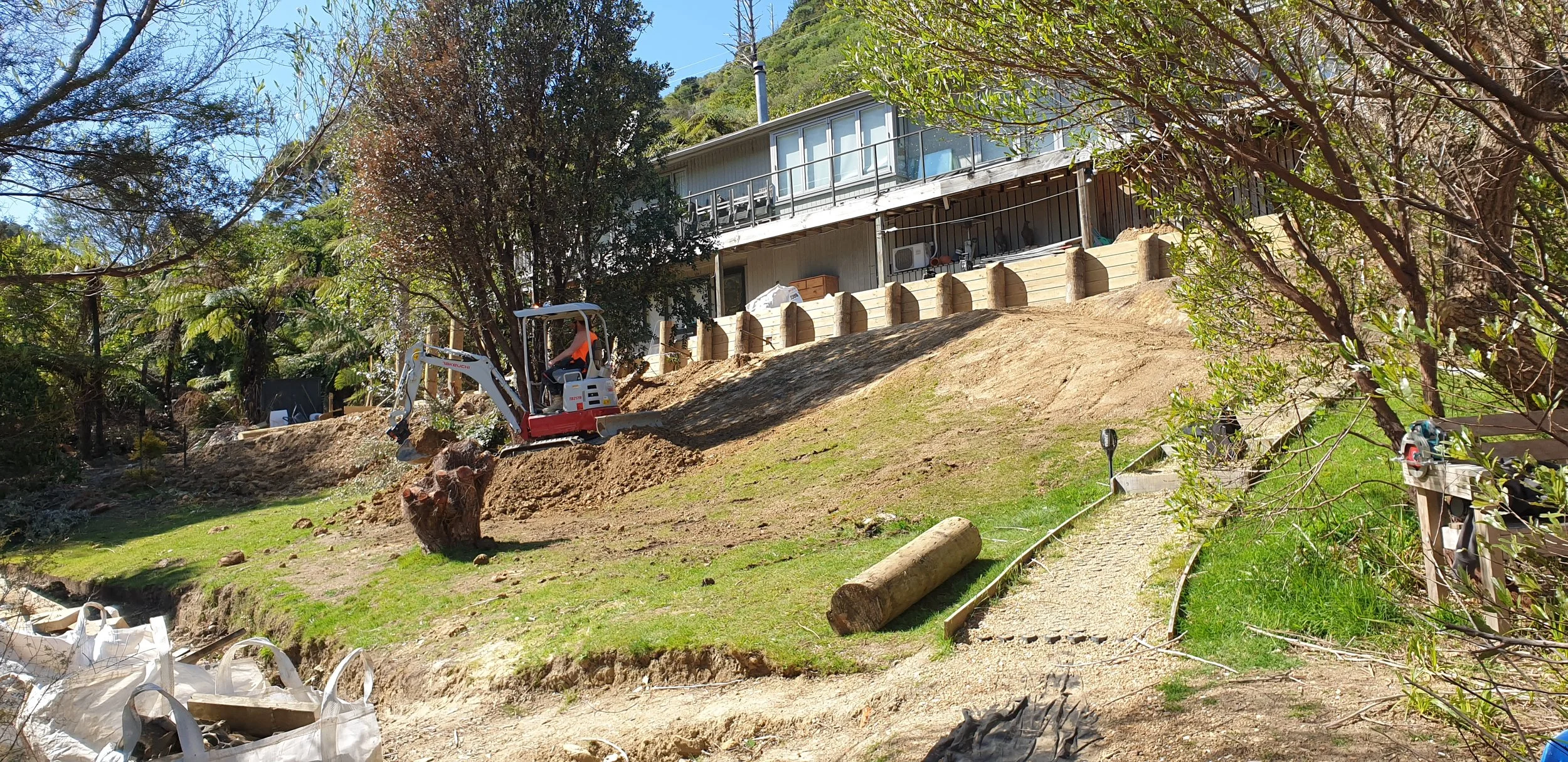 A backyard with a house on a hill, construction work in progress with a mini excavator, soil moves, and trees around.