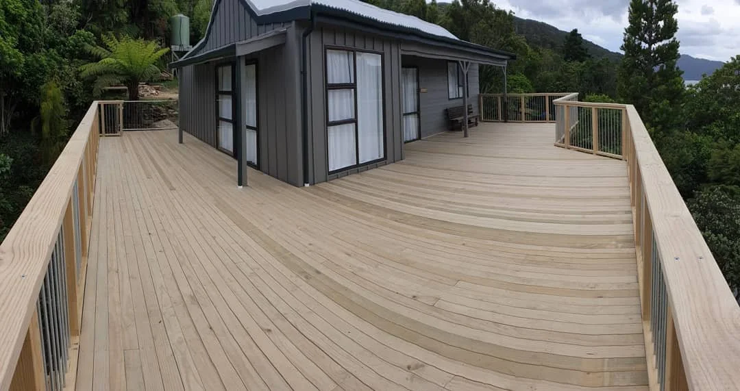 A large wooden deck attached to a gray house surrounded by trees and hills.