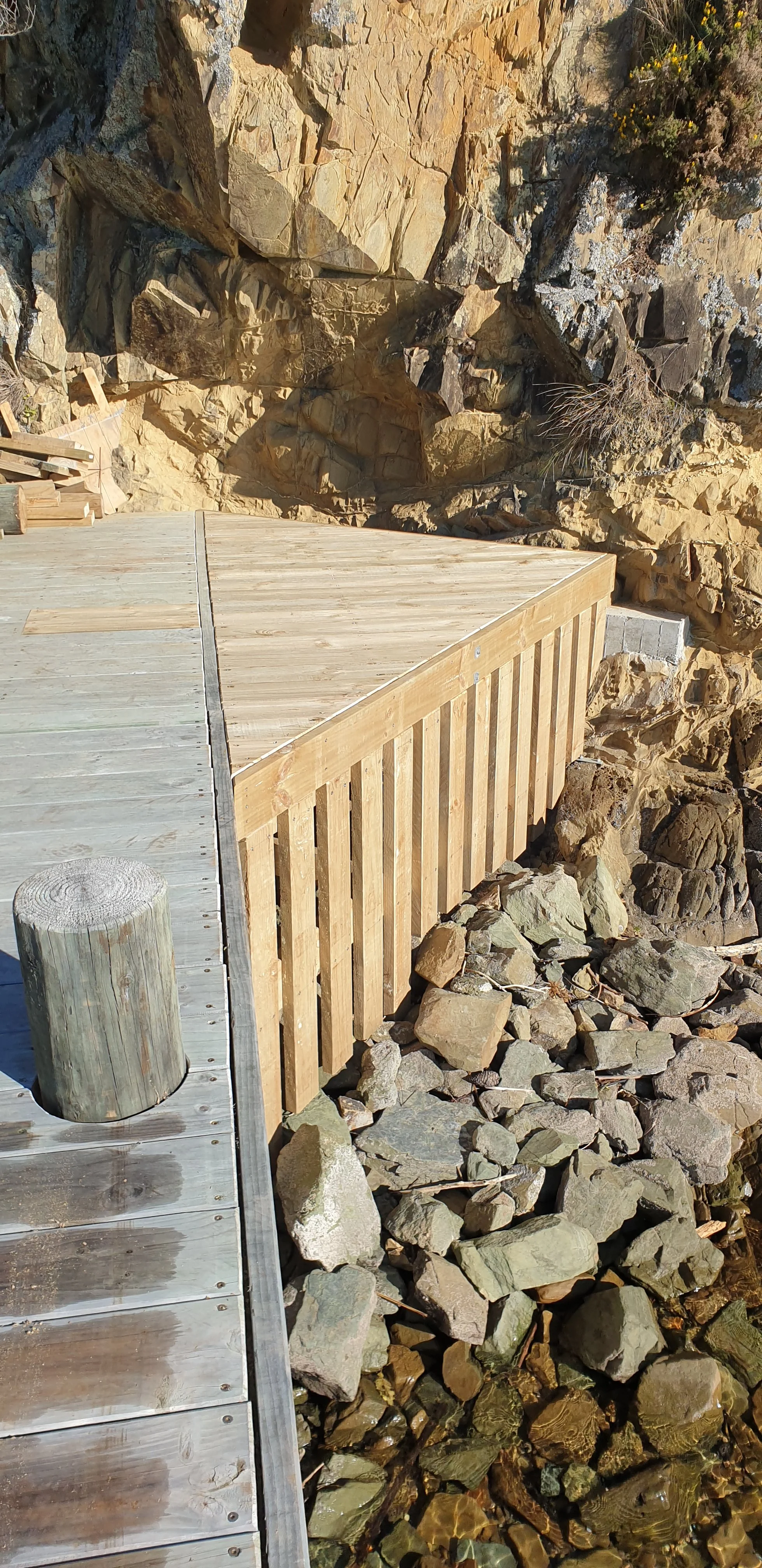 A wooden dock extending over rocky water, with a rocky cliff in the background.