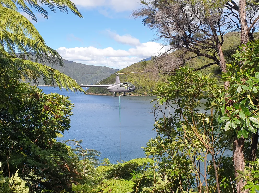 A helicopter hovering above a lake surrounded by green trees and hills under a partly cloudy sky.