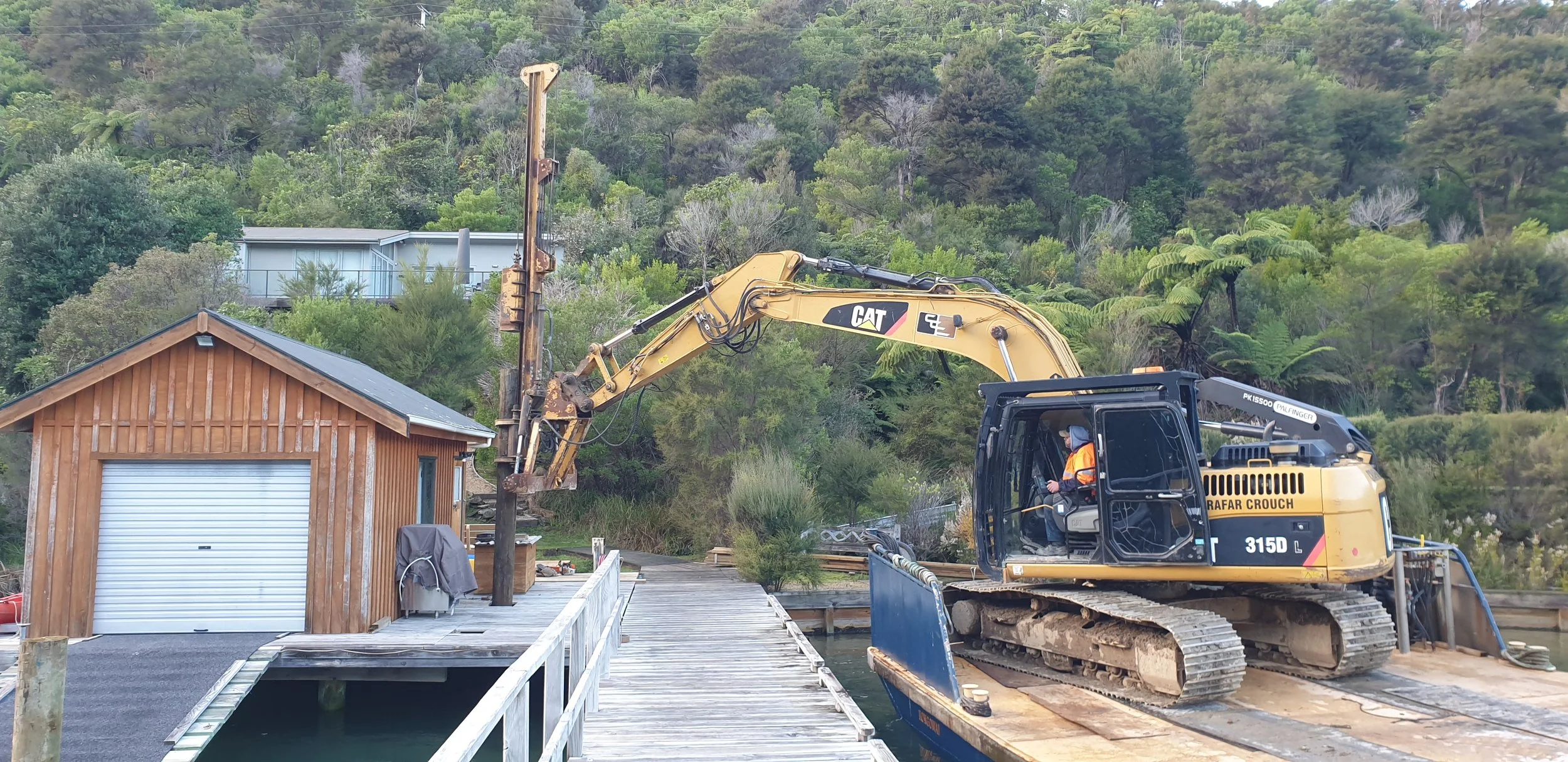 A construction excavator on a wooden dock next to a small wooden building with a white garage door, surrounded by trees and greenery on a hillside.
