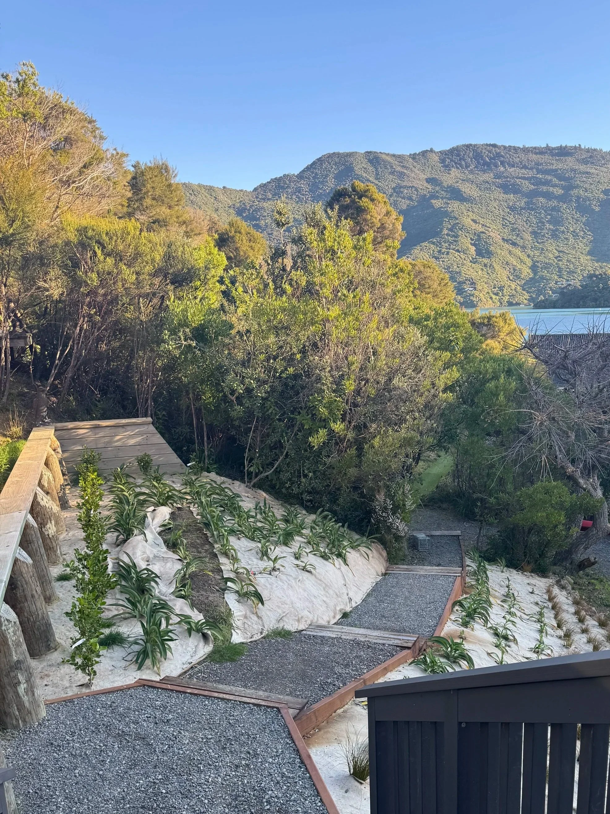 A scenic view of a landscaped garden with a gravel pathway, green bushes, and trees, overlooking a body of water with hills in the background under a clear blue sky.