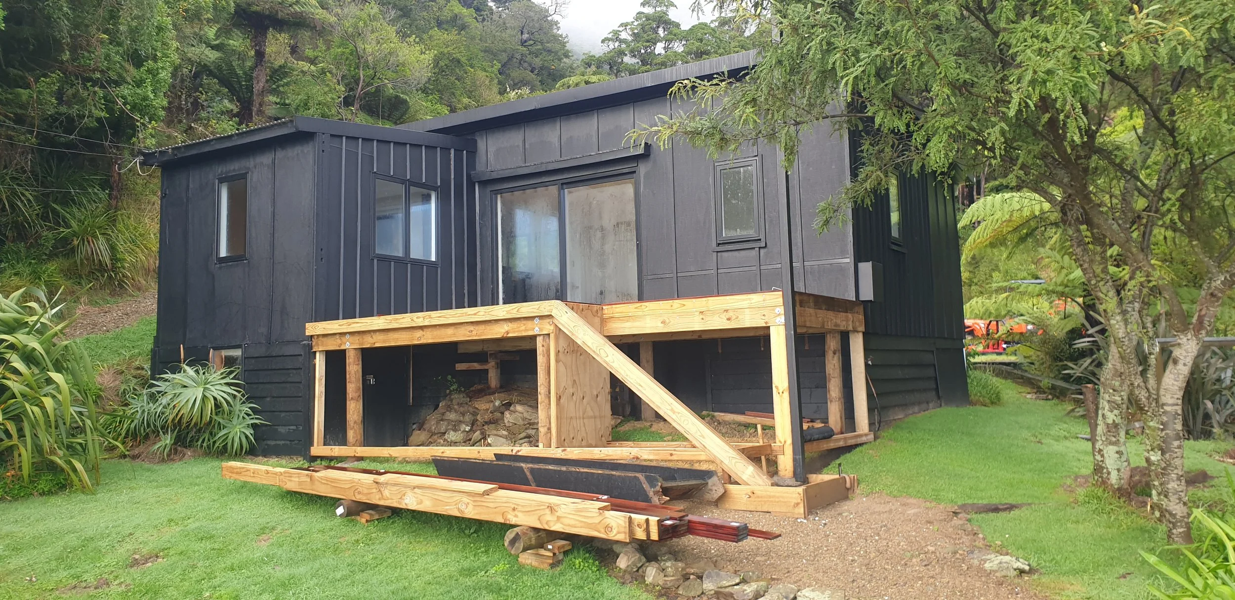 A black house under construction with a wooden porch and a grassy yard, surrounded by trees and plants.