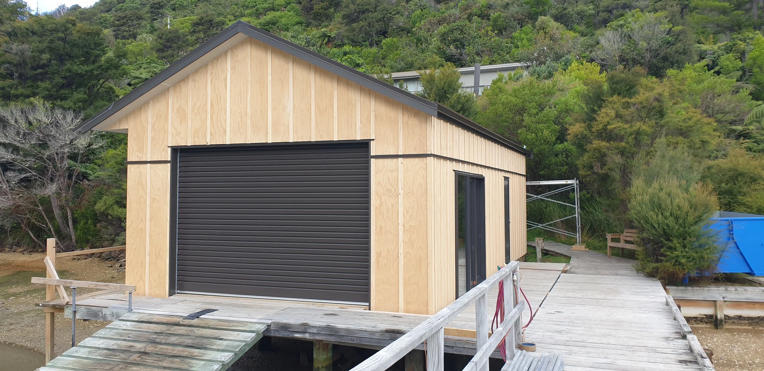 A partially constructed wooden building with a black roll-up garage door, situated on a wooden deck in a green, wooded area. There is scaffold on the side and some construction materials around.