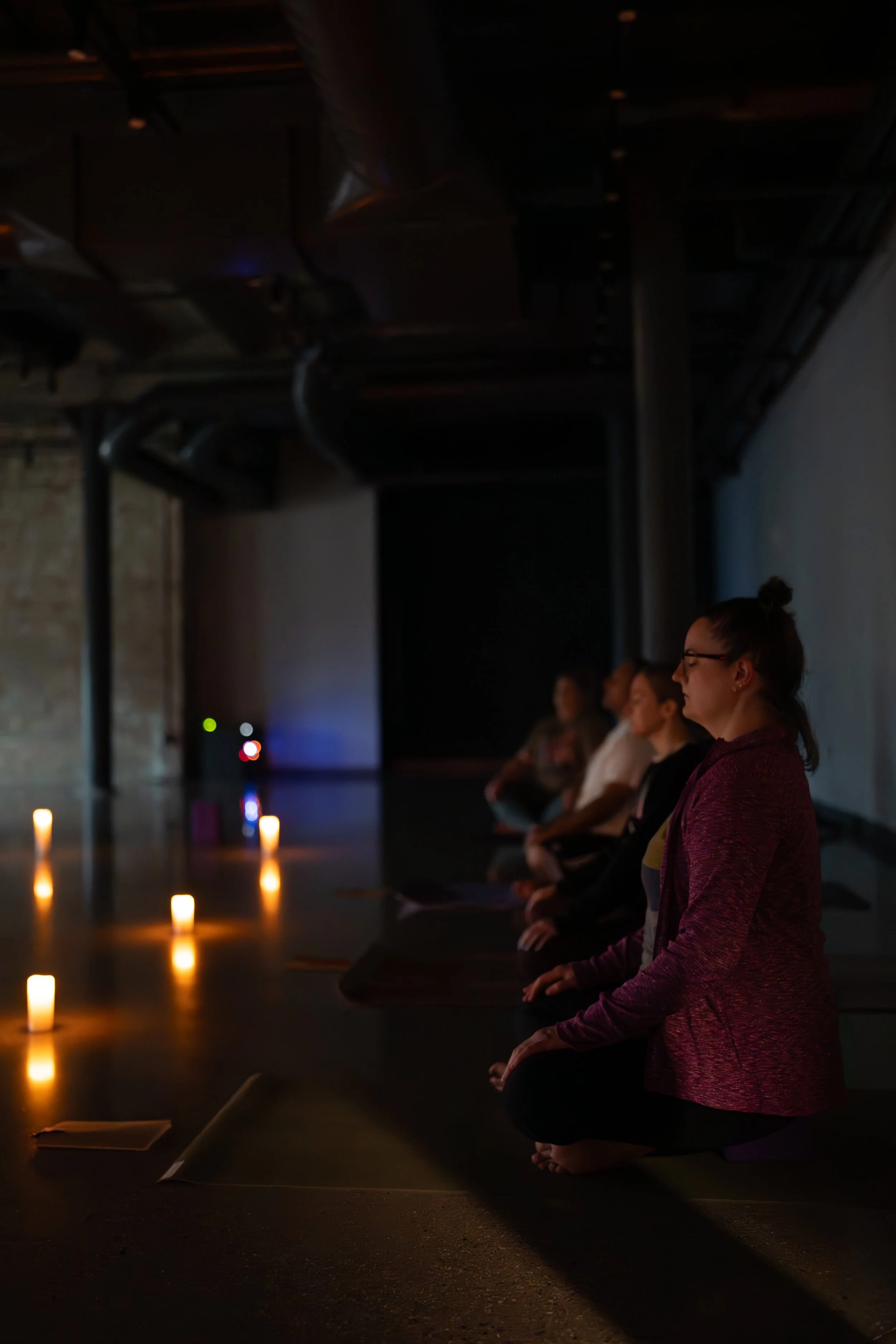 Group of women practicing yoga or meditation in a spacious, well-lit indoor studio with yoga mats, water bottles, and candles.