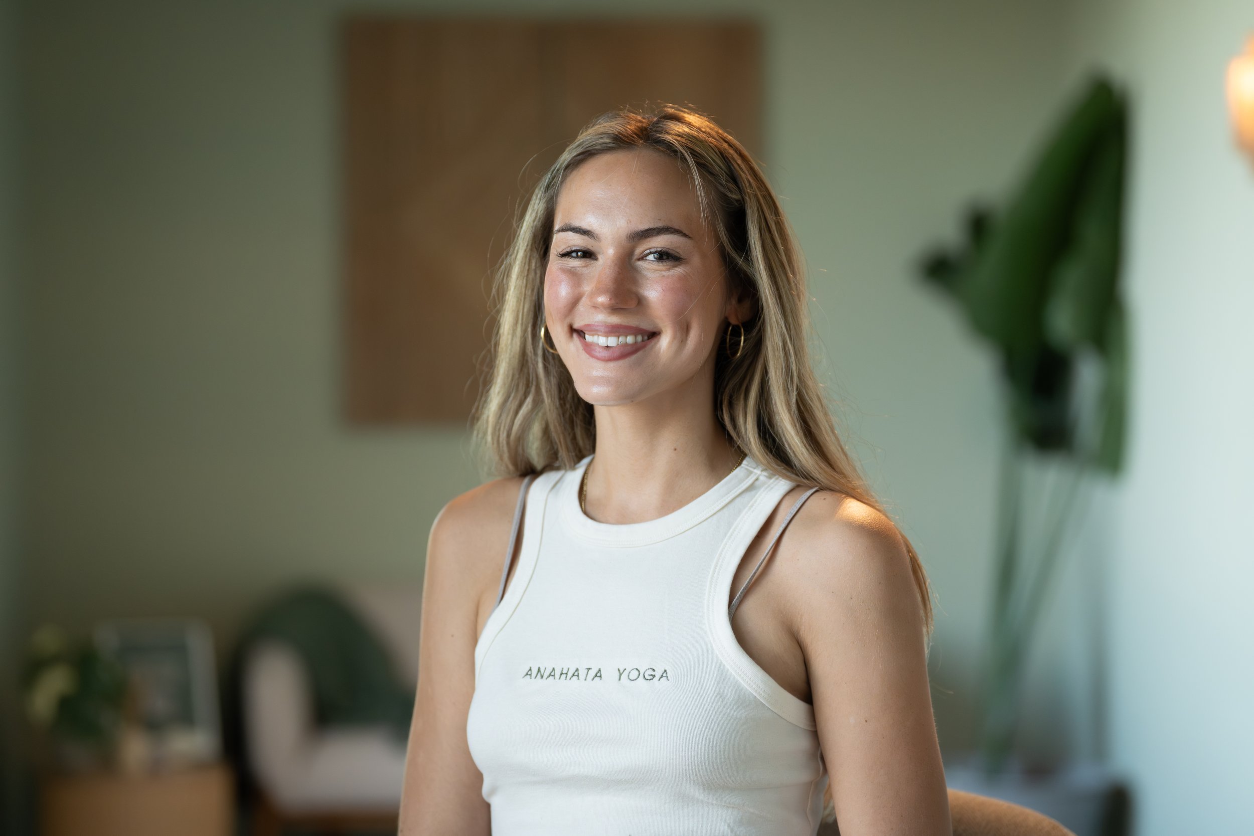 Young woman with long wavy hair sitting on the floor in front of a plain white backdrop, smiling softly, with her hands on the ground and legs crossed, dressed in a black top, black pants, white socks, and sneakers.