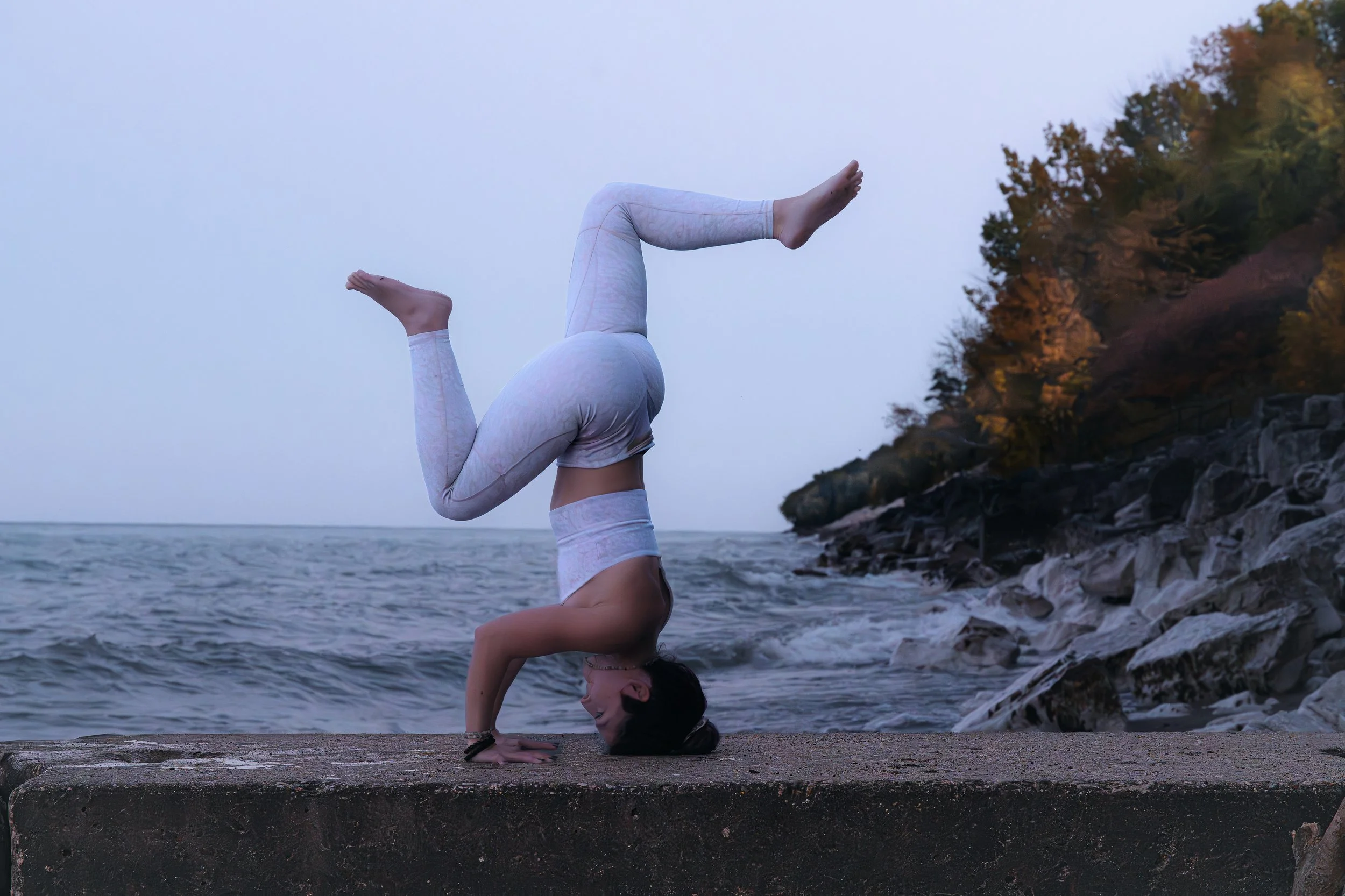 A woman in white workout clothes performing a headstand on a concrete surface near the seaside with trees on a hillside in the background.