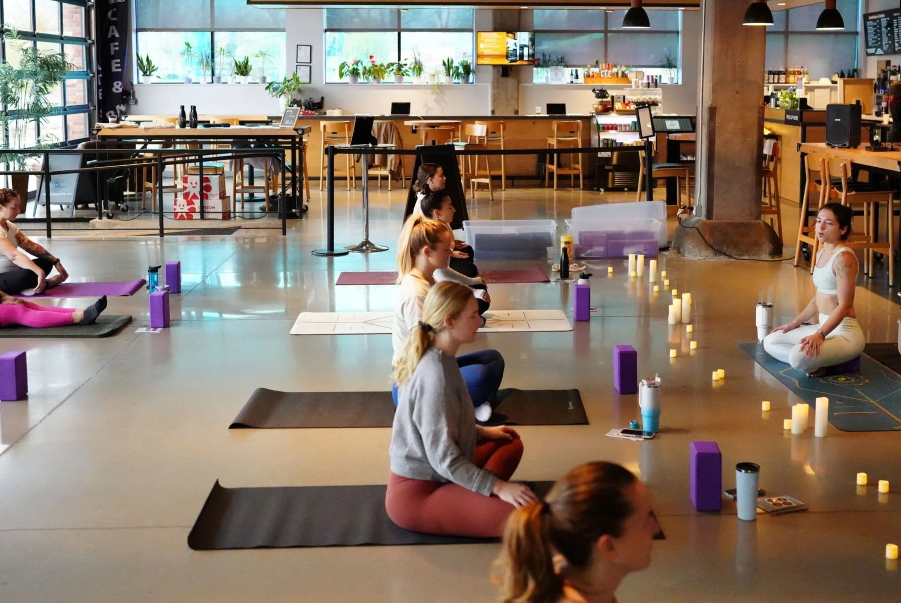 Group of women practicing yoga or meditation in a spacious, well-lit indoor studio with yoga mats, water bottles, and candles.