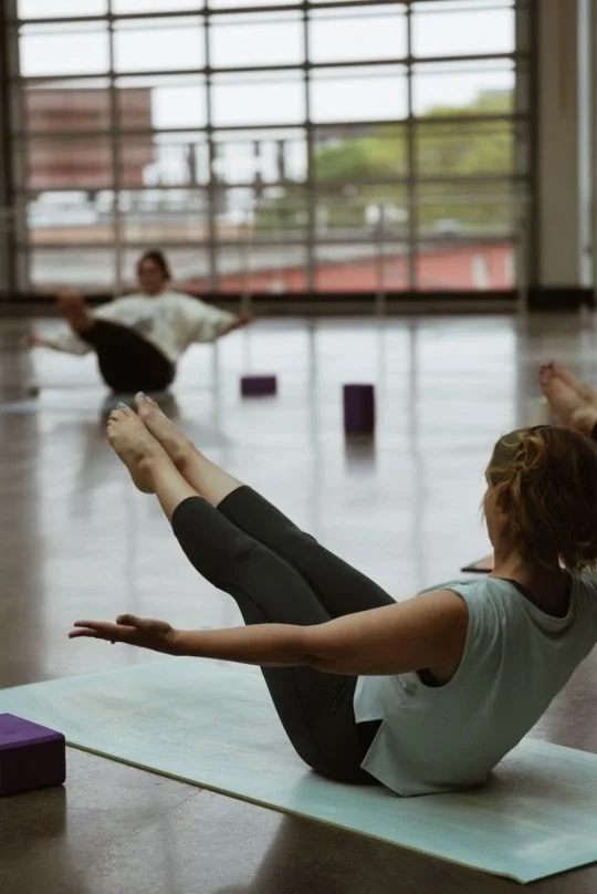 A girl doing yoga on a mat in a spacious indoor studio with large windows. In the background, a child is sitting on the floor with legs extended and arms out.