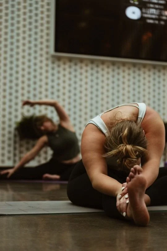 Two women practicing yoga in a studio, one in foreground doing seated forward bend, and one in background doing a side stretch on a yoga mat.