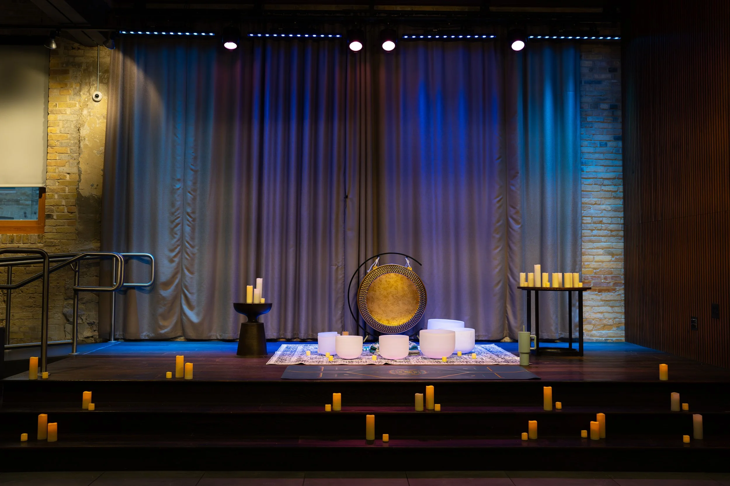 Empty stage with silver curtains, candles, white singing bowls, a gong, and a rug, decorated for a performance or ceremony, with yellow candles on steps in front.