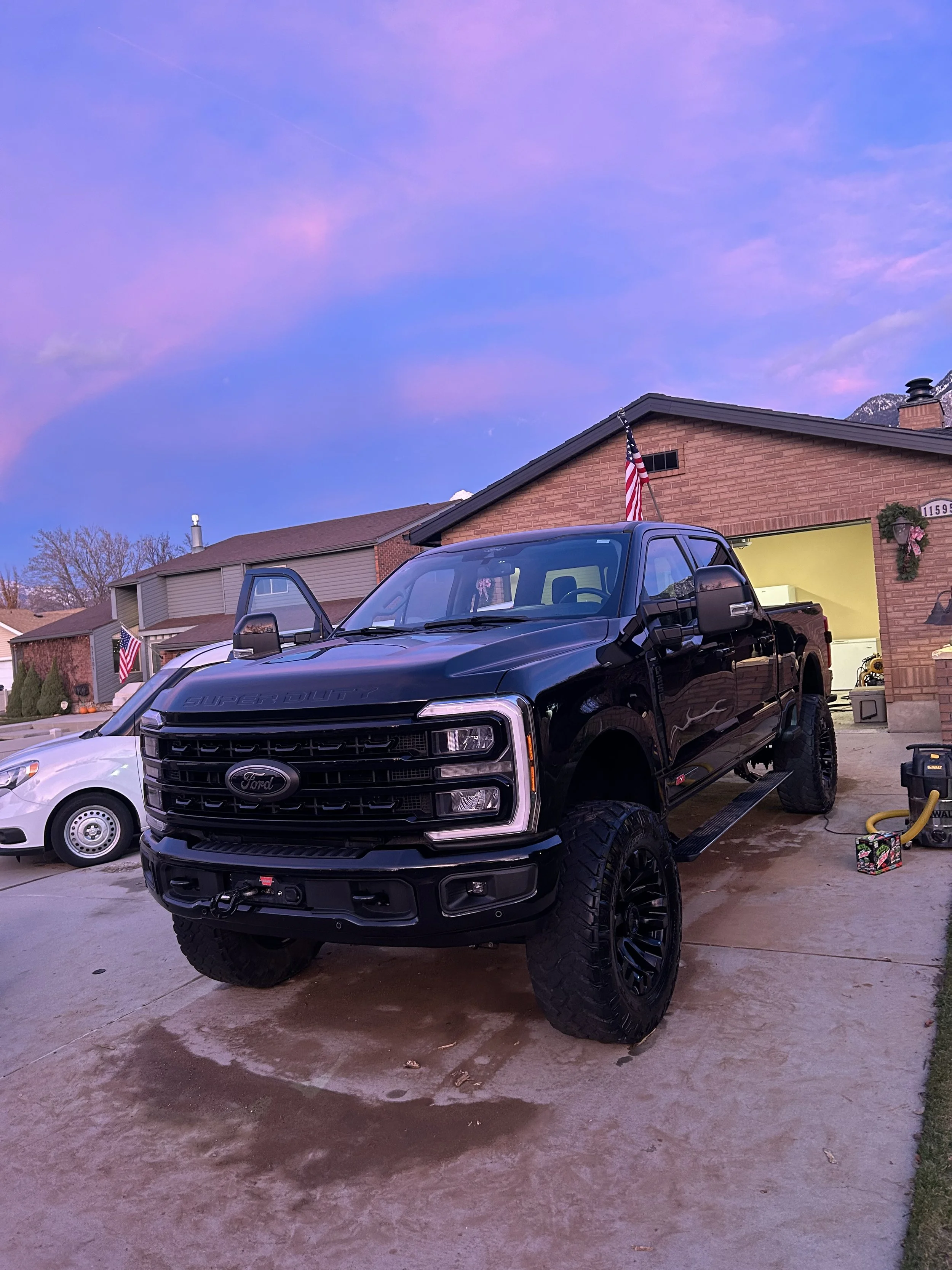 Black Ford Super Duty truck parked in a residential driveway at sunset with a purple sky, next to a white car, with American flags and a house in the background.
