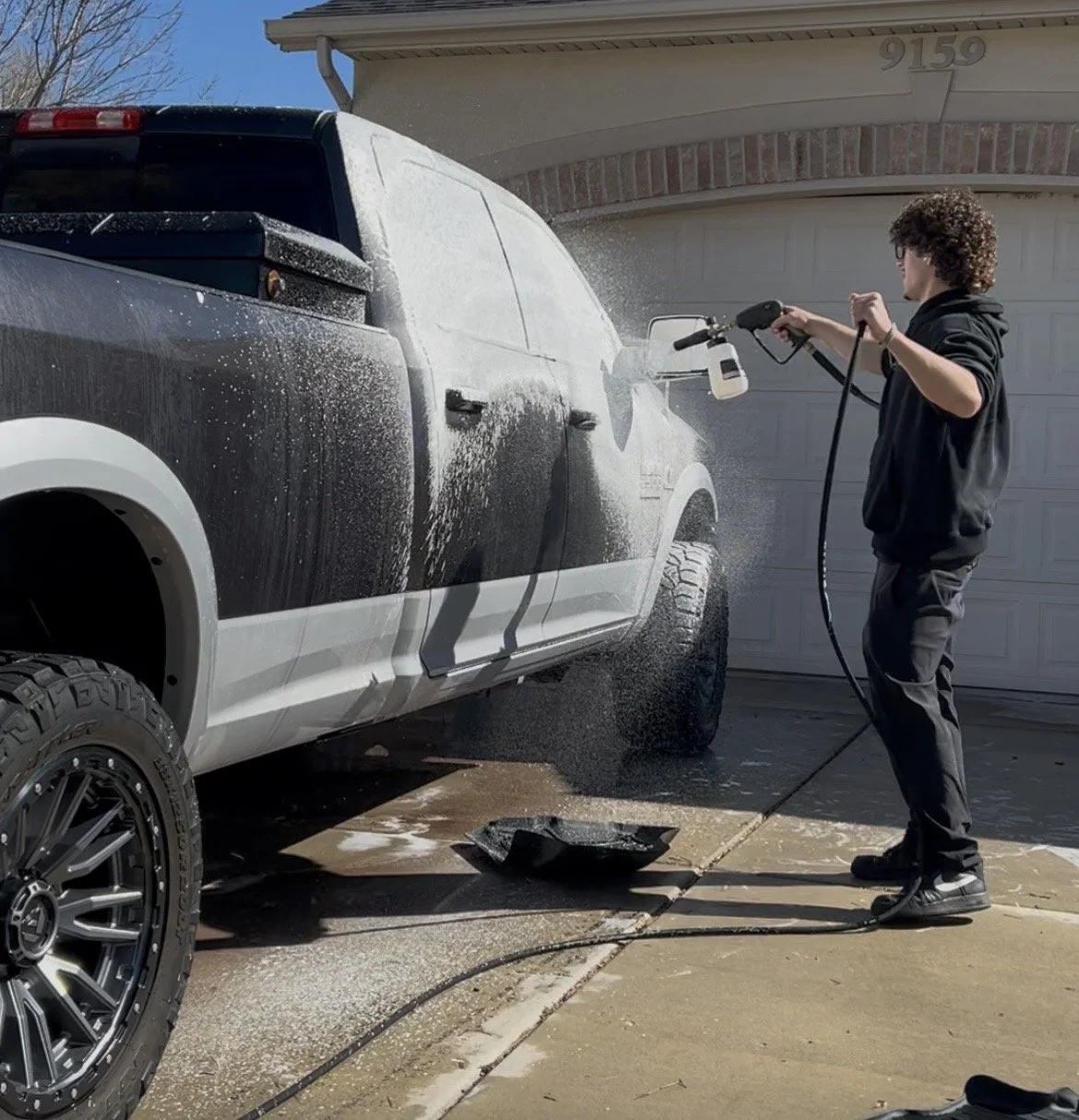 Person using a power washer to clean a white and black pickup truck parked outside a house.