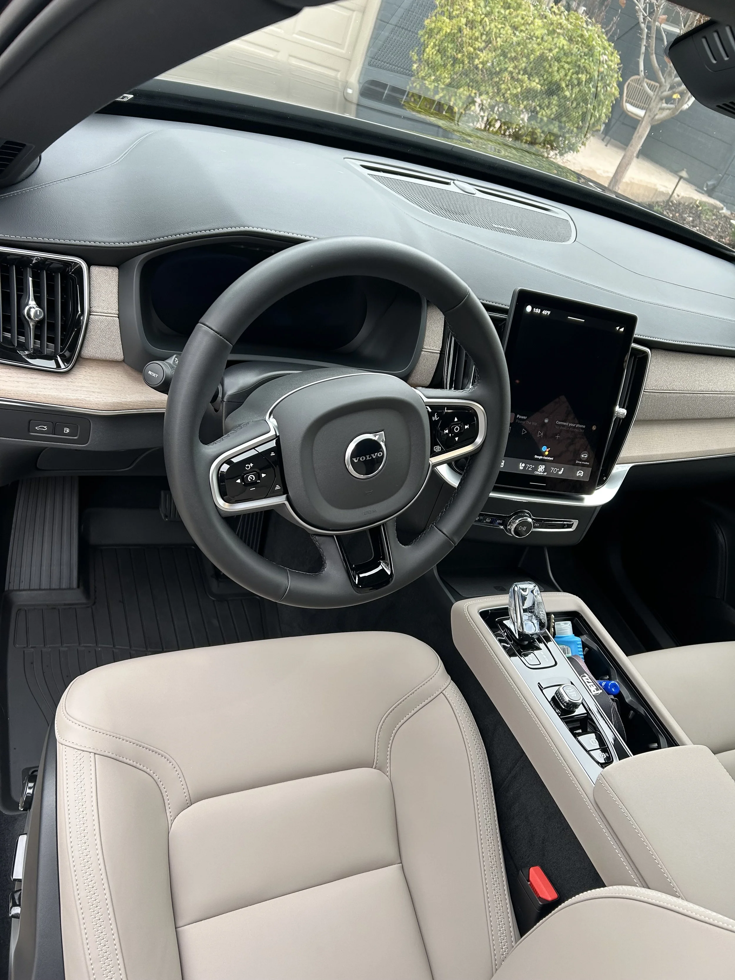 Interior of a modern Volvo car, showing a beige seat, steering wheel with Volvo logo, large touchscreen display, gear shift, and dashboard.
