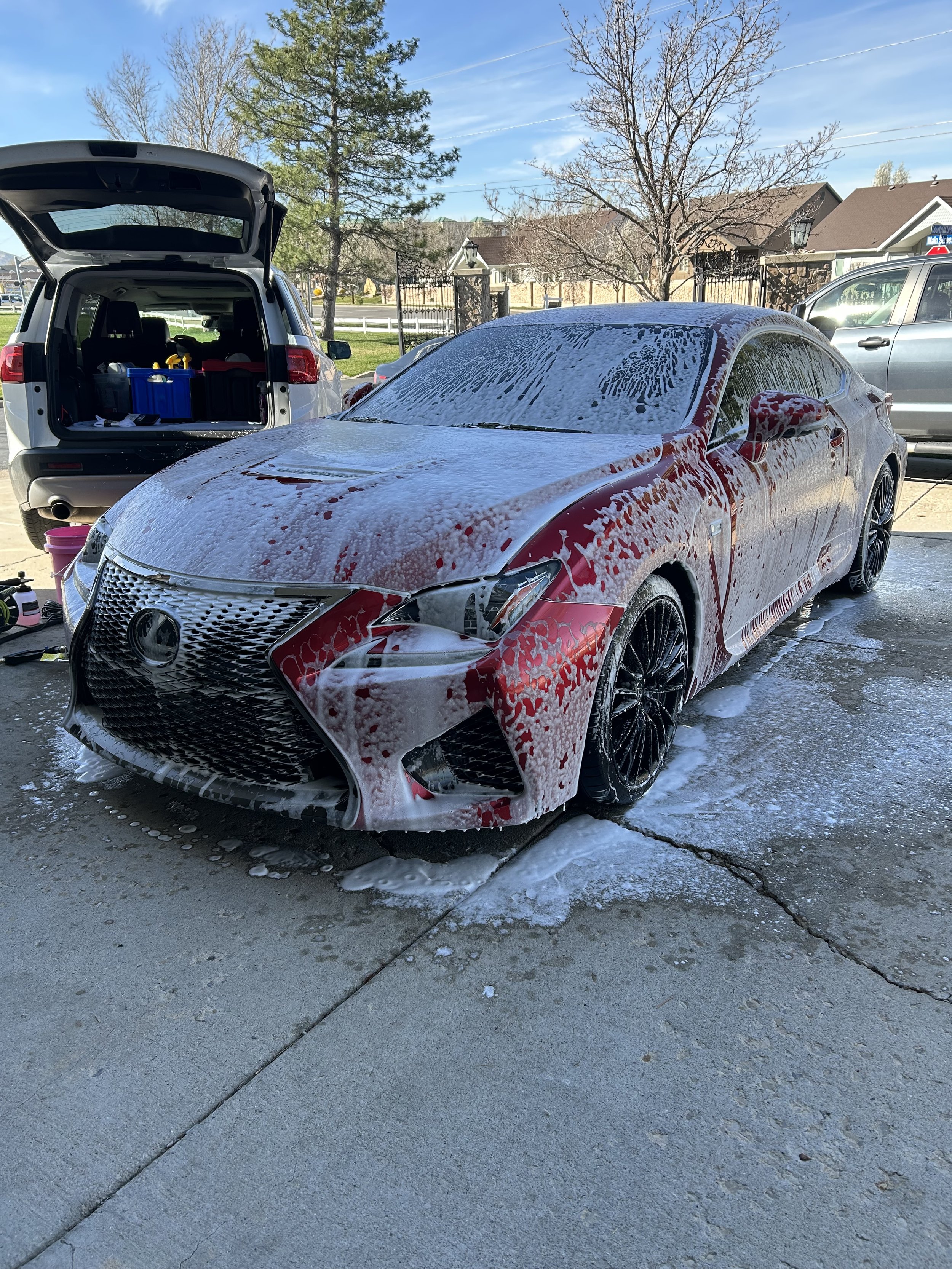 A red car being washed with soap on the driveway, foam covering the car's surface, next to an open car trunk with cleaning supplies inside, in a suburban neighborhood.