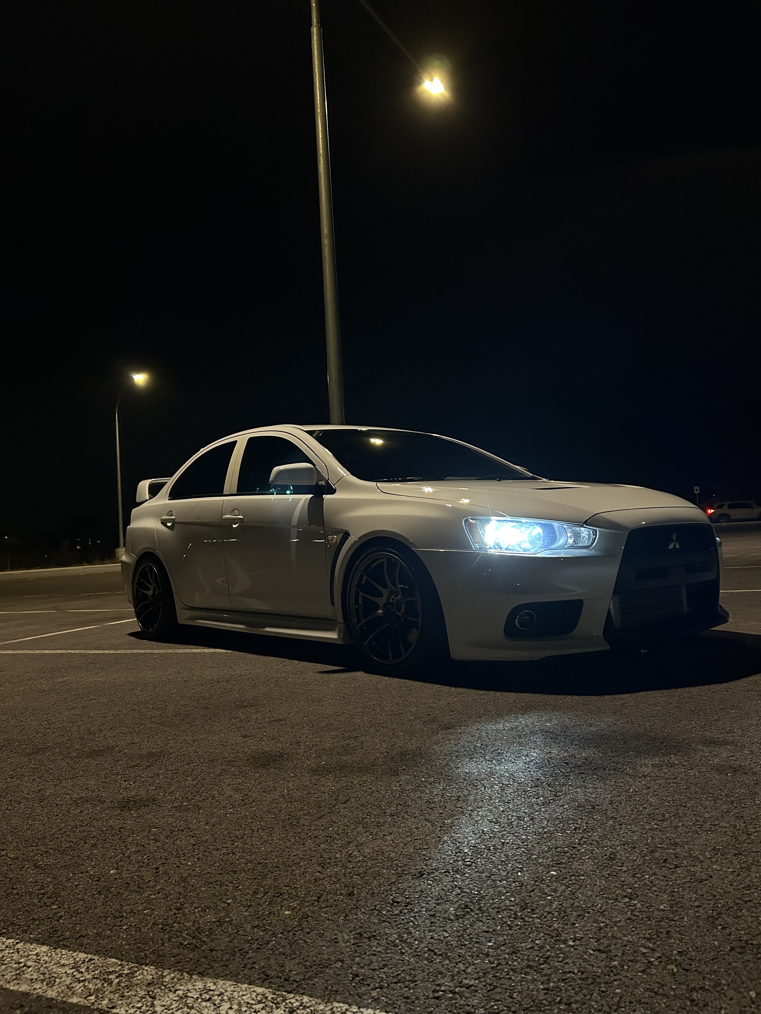 A silver Mitsubishi Lancer Evolution parked at night in an empty parking lot with streetlights illuminating the scene.