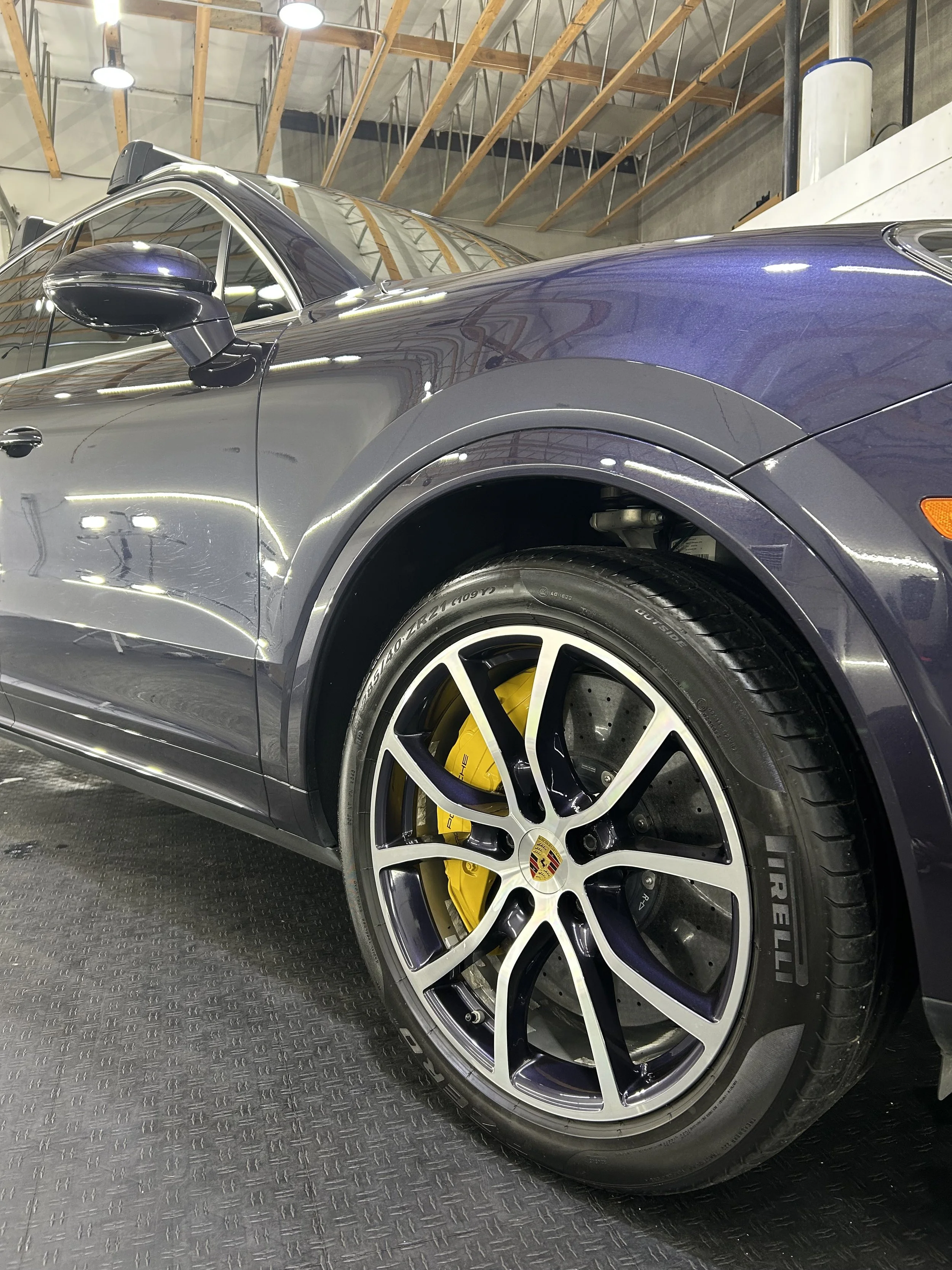Close-up of a dark gray Porsche sports car showing the front left wheel with a yellow brake caliper and black tire, inside a garage or showroom with an unfinished ceiling.