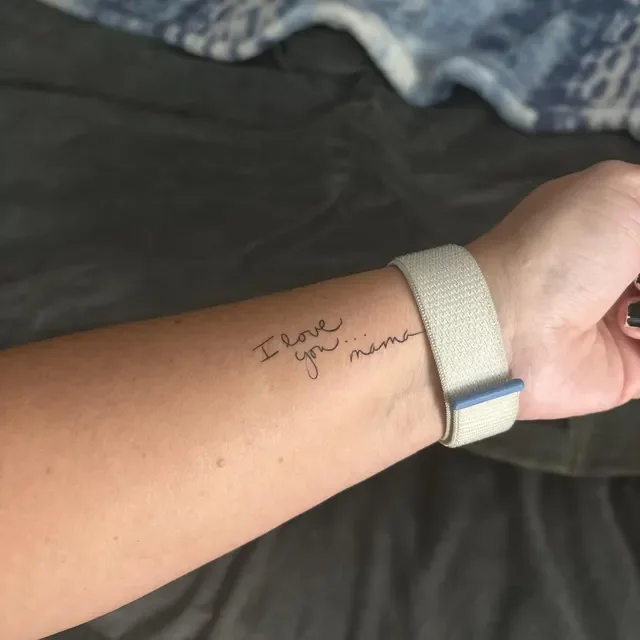 A person's forearm with a handwritten tattoo that says 'I love you... mama'. The person is wearing a beige wristband and has dark nail polish.