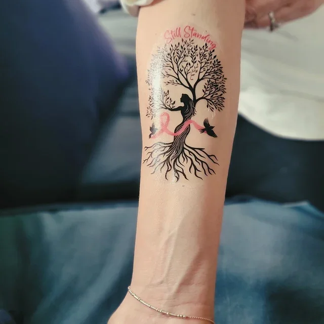 Tattoo of a tree with a woman sitting on the trunk, pink ribbon wrapping around it, birds flying nearby, and the words "Still Standing" above the tree.