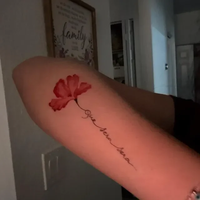 A tattoo of a red flower with black stems and the cursive words "You be my flower" on a person's forearm.