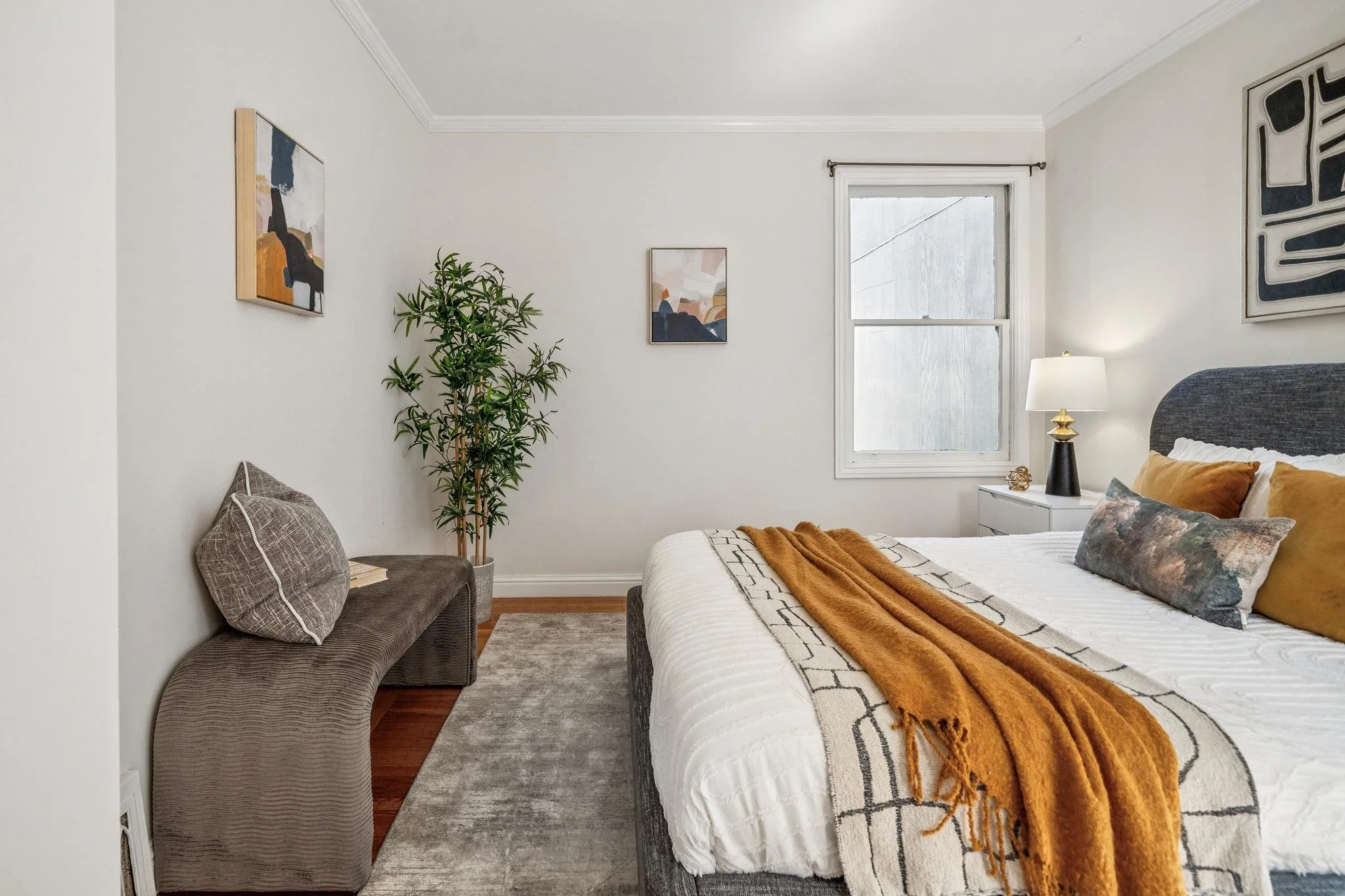A bedroom with white walls, a grey upholstered bed with various pillows and an orange throw blanket, a nightstand with a lamp, a window with rain on the glass, a grey curved bench with cushions, a potted plant, and framed abstract artwork on the wall