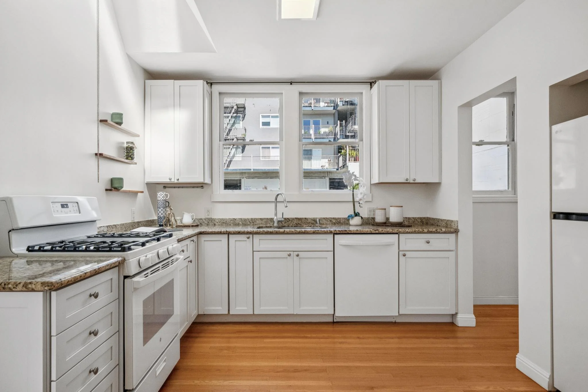 Clean white kitchen with granite countertops, white cabinets, and hardwood floors. A window over the sink shows a view of neighboring buildings.