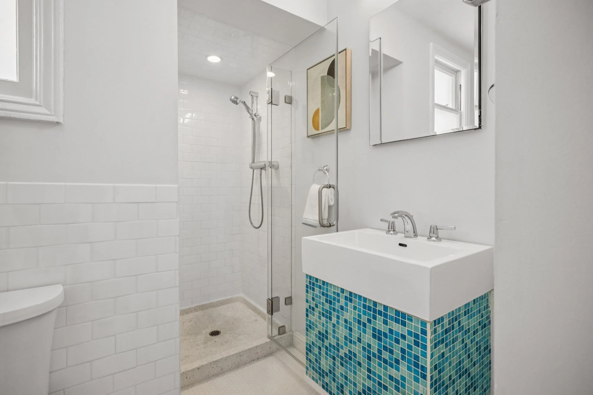 Modern bathroom with glass-enclosed walk-in shower, white subway tiles, small mosaic blue-tiled vanity, white walls, mirror, window, and abstract art on the wall.
