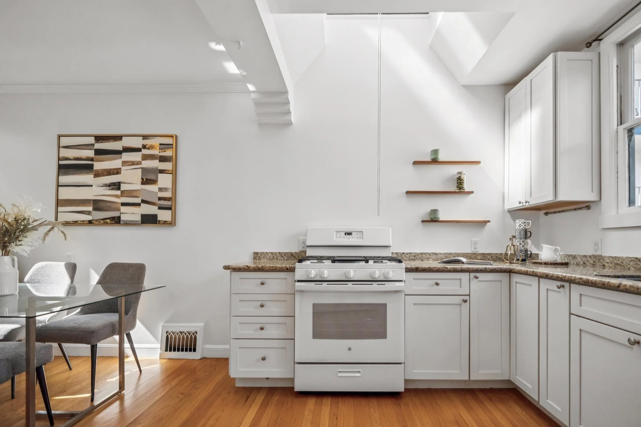 A bright and minimalistic kitchen with white cabinets, a granite countertop, a white stove, and a window on the right. There's a dining table with four chairs on the left, a framed abstract artwork on the wall, and three small wooden shelves with dec