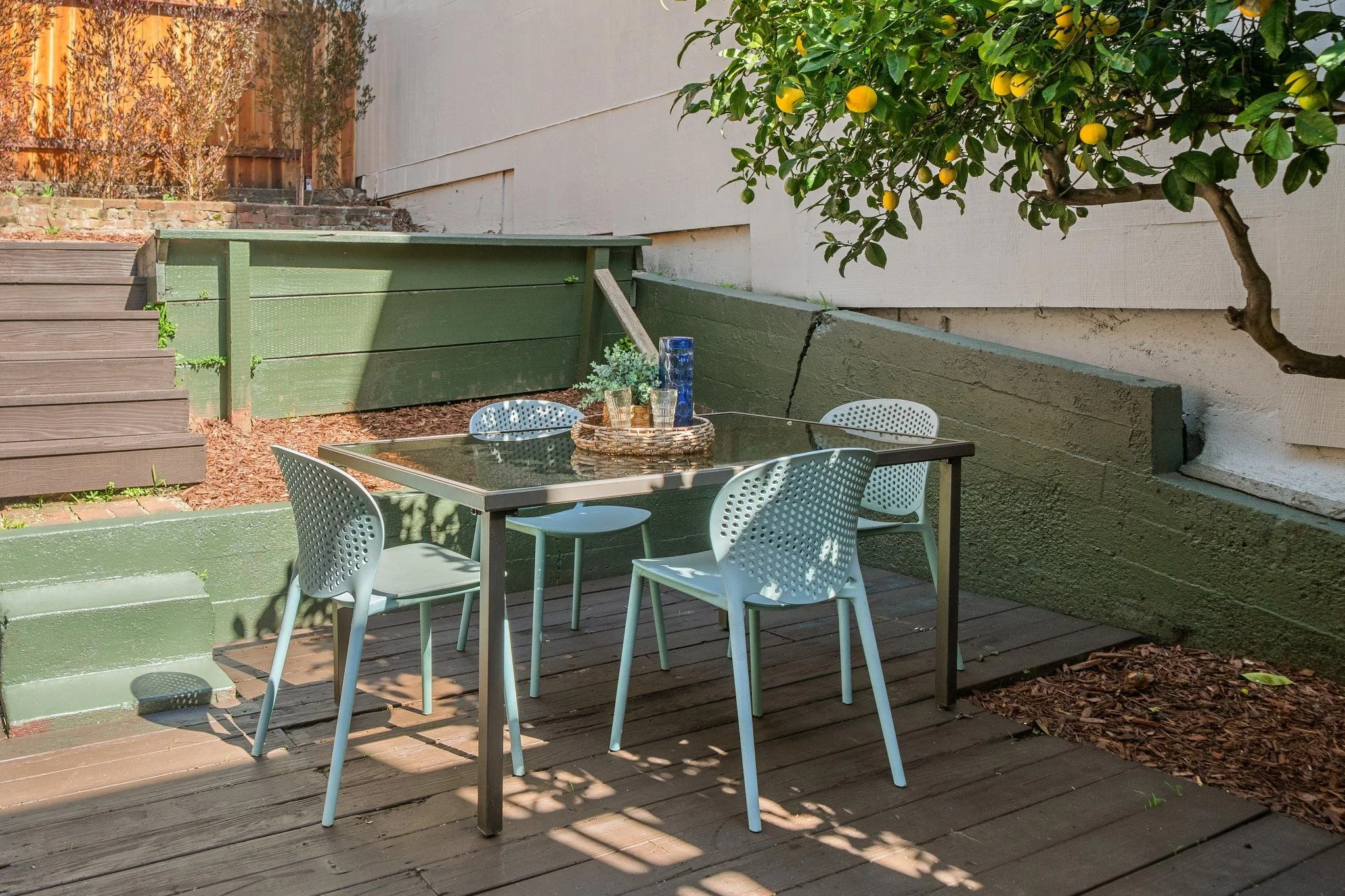 An outdoor patio with a glass-top table and four light blue chairs. There is a tray with glassware on the table, and a lemon tree with yellow fruit growing nearby. The patio has wooden decking and a green painted block wall, with stairs leading up on