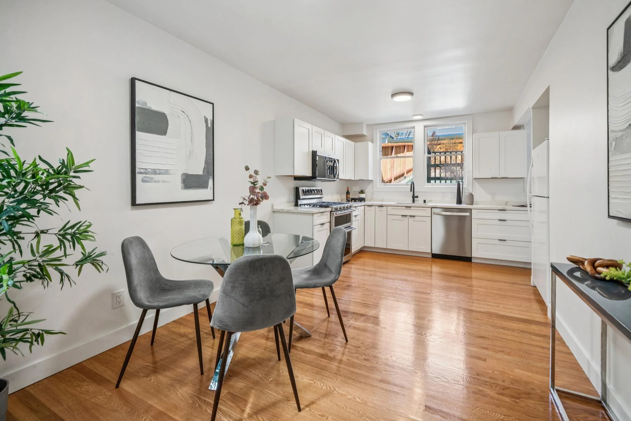 Modern white kitchen and dining area with hardwood floors, a round glass table with four gray chairs, and minimal decoration.