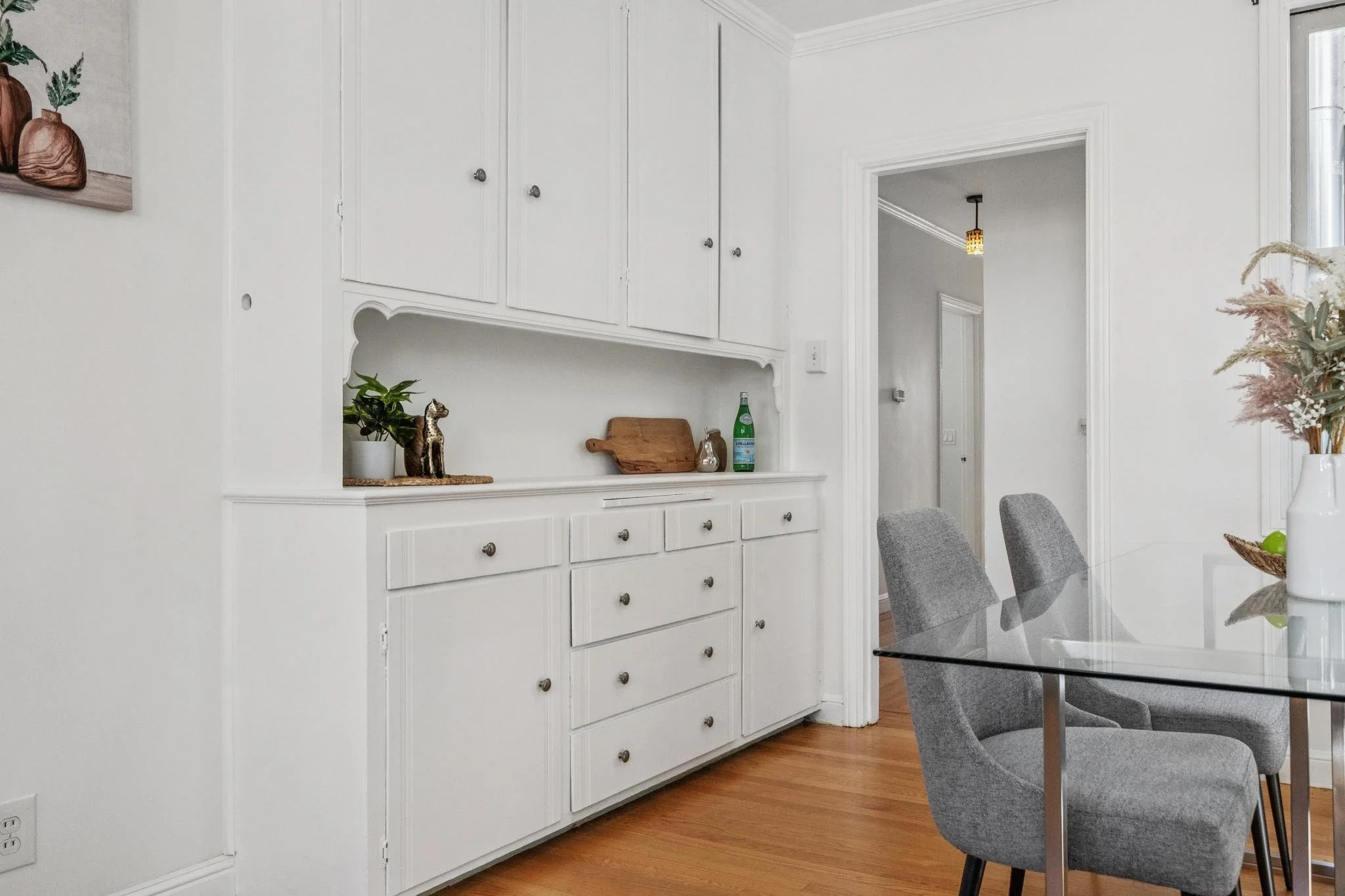 A dining room with white built-in cabinets and a glass-top table with gray chairs. There are decorative items on the cabinet and a vase with dried flowers on the table.