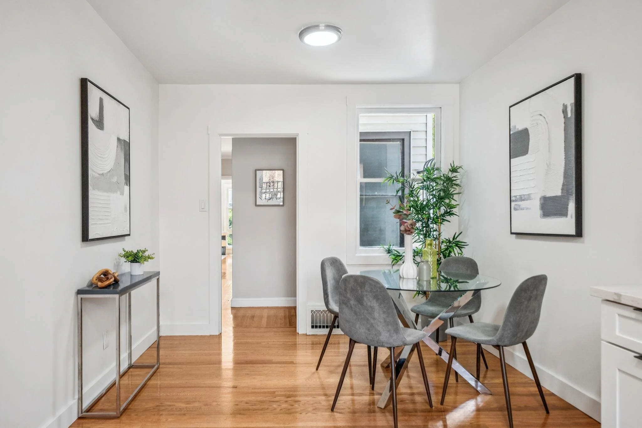 Modern dining area with a glass table, gray upholstered chairs, and floral arrangements, with black and white abstract artwork on white walls and a hardwood floor.