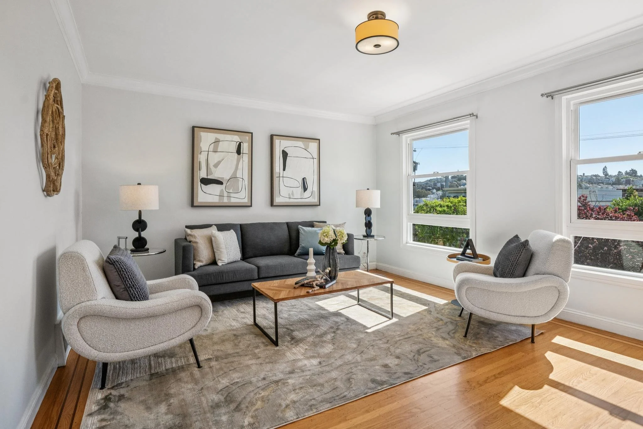 Bright living room with white walls, hardwood floors, and large windows. Contains a gray sofa, two white armchairs, a wooden coffee table, and two side tables with lamps. Wall art and decor accents add to the modern decor.
