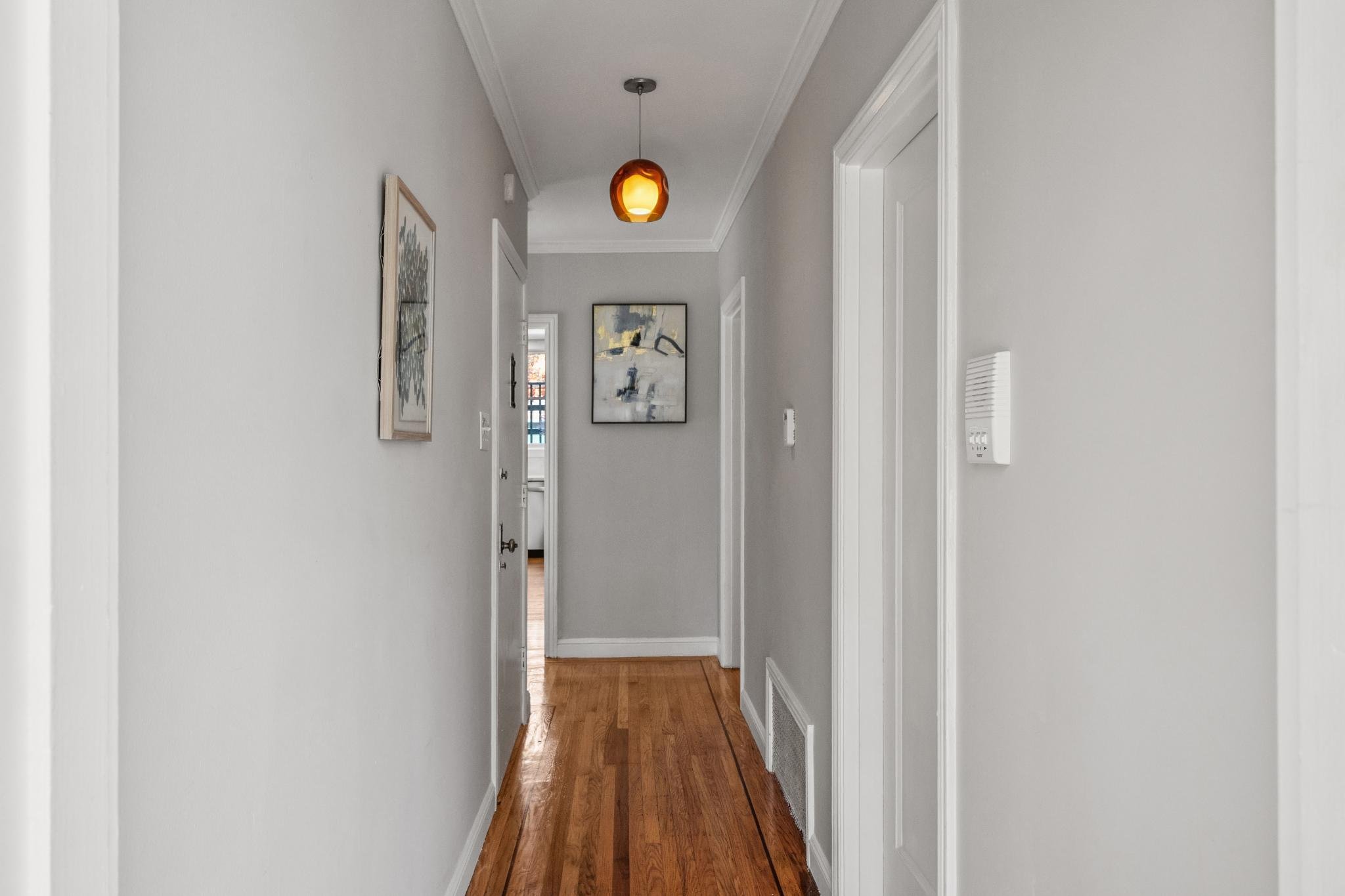 A narrow hallway with light gray walls, wooden flooring, and white door frames. There are two abstract paintings on the walls and a hanging light fixture with an orange shade.