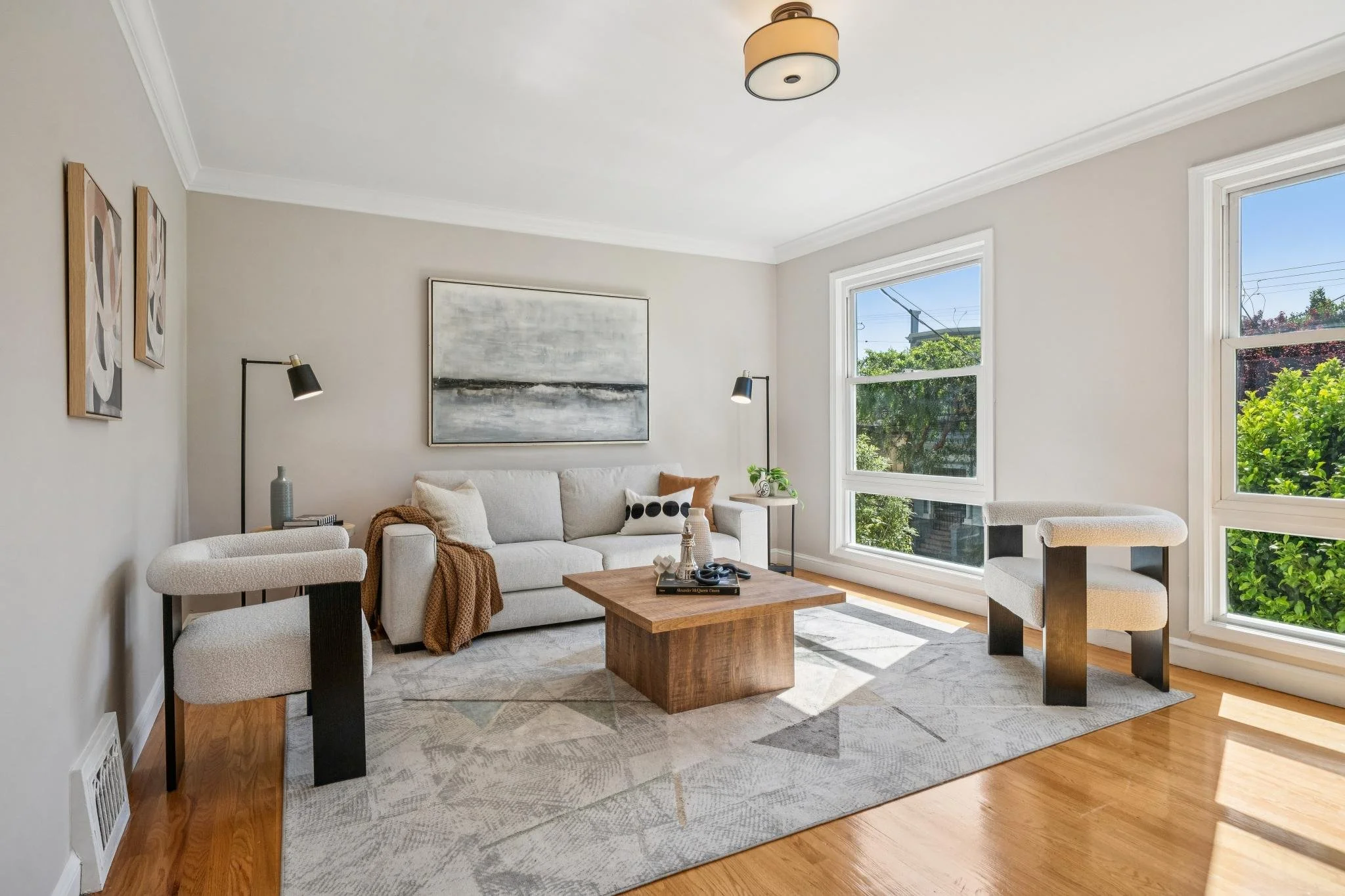 Bright living room with beige walls, large windows, a white sofa with pillows, two modern armchairs, a wooden coffee table, abstract wall art, and a ceiling light fixture.