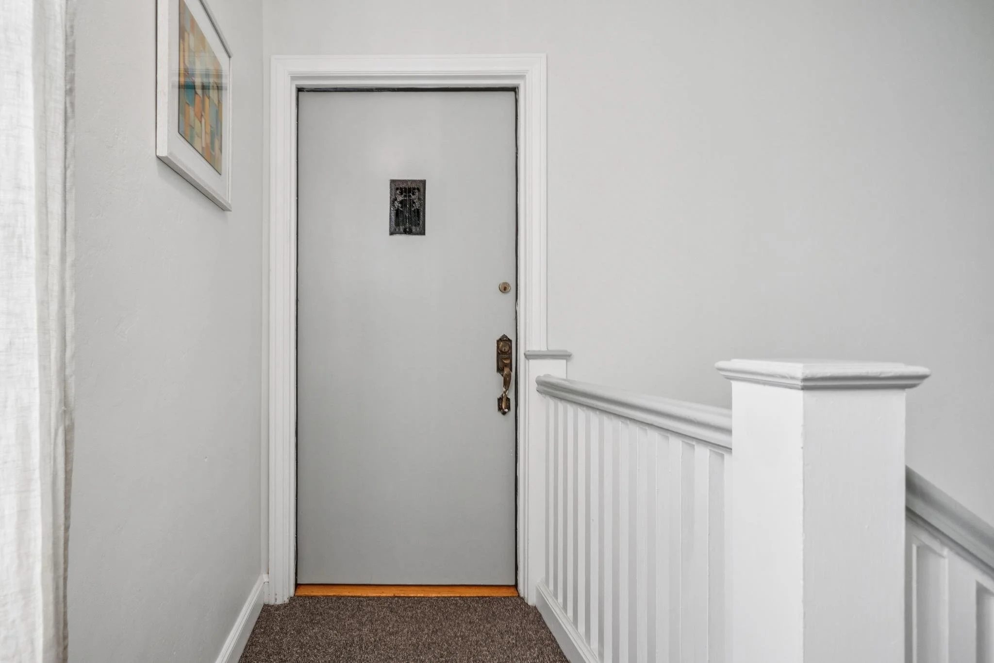 Interior view of a white hallway leading to a gray door with a small window. The hallway has white walls, a framed picture hanging on the left wall, and a partial white wooden railing on the right. The floor has a textured brown carpet.