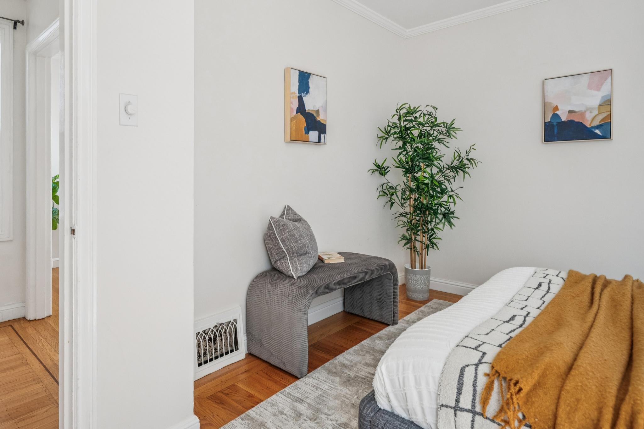 A bedroom corner with a bed, a gray bench with pillows, two abstract paintings on a white wall, a large potted plant, hardwood floors, and a beige decorative rug.