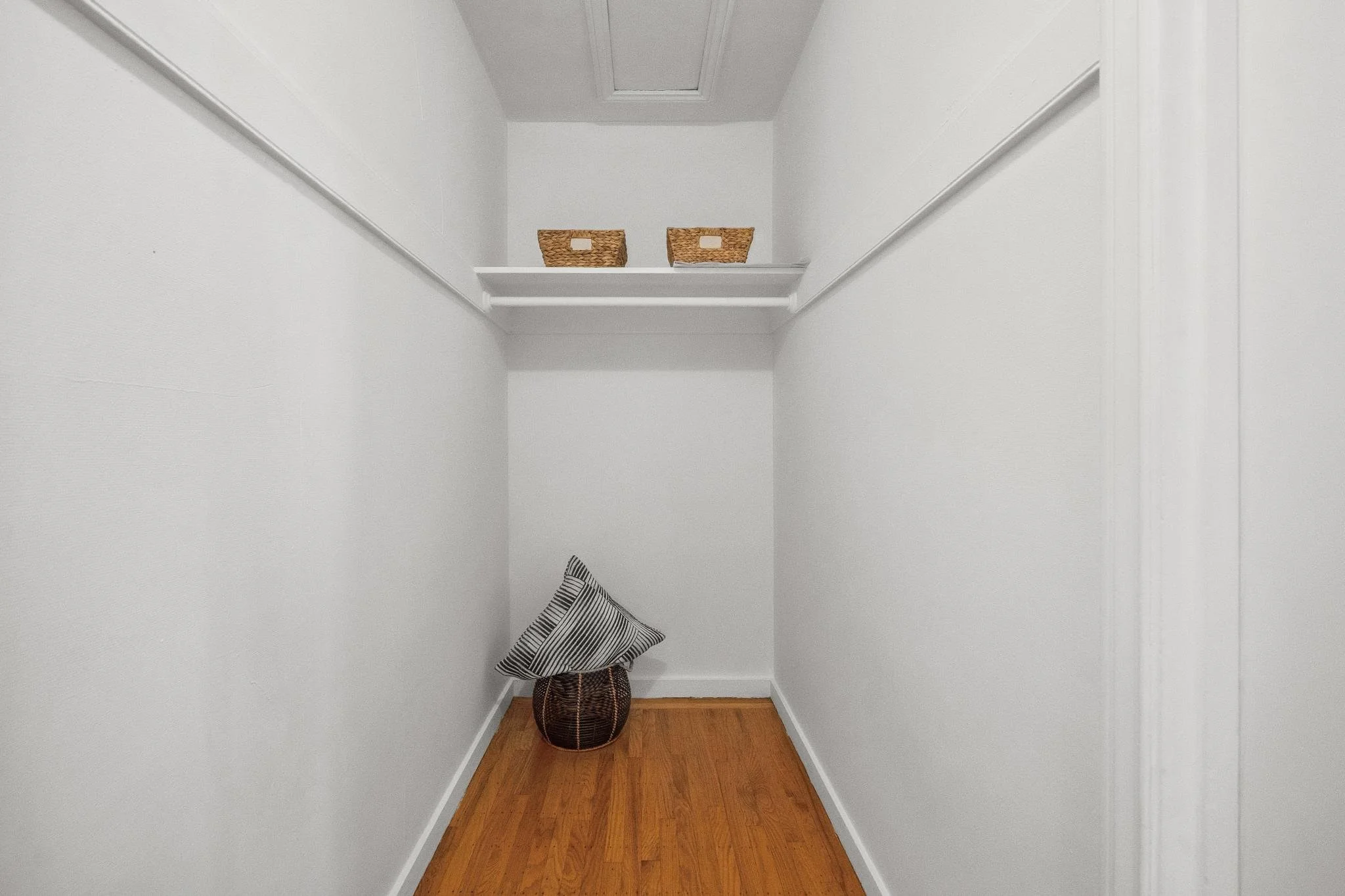 Empty walk-in closet with white walls, a small shelf with two wicker baskets, a floor basket with a striped pillow, and a ceiling vent.
