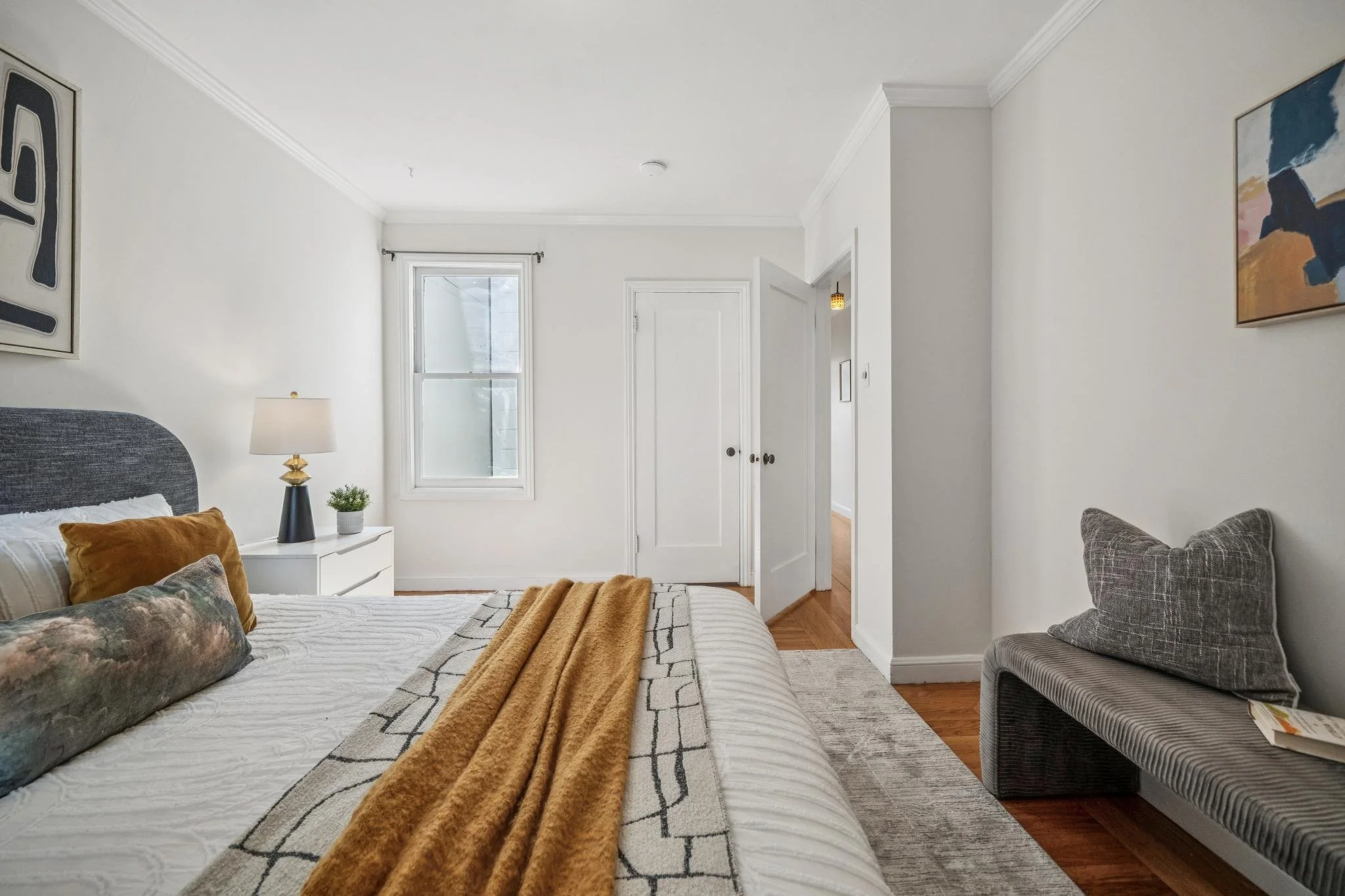 A bedroom with a bed, a lamp, a small plant, and artwork on the walls. There is a window, a door, and a gray bench with a plaid pillow.