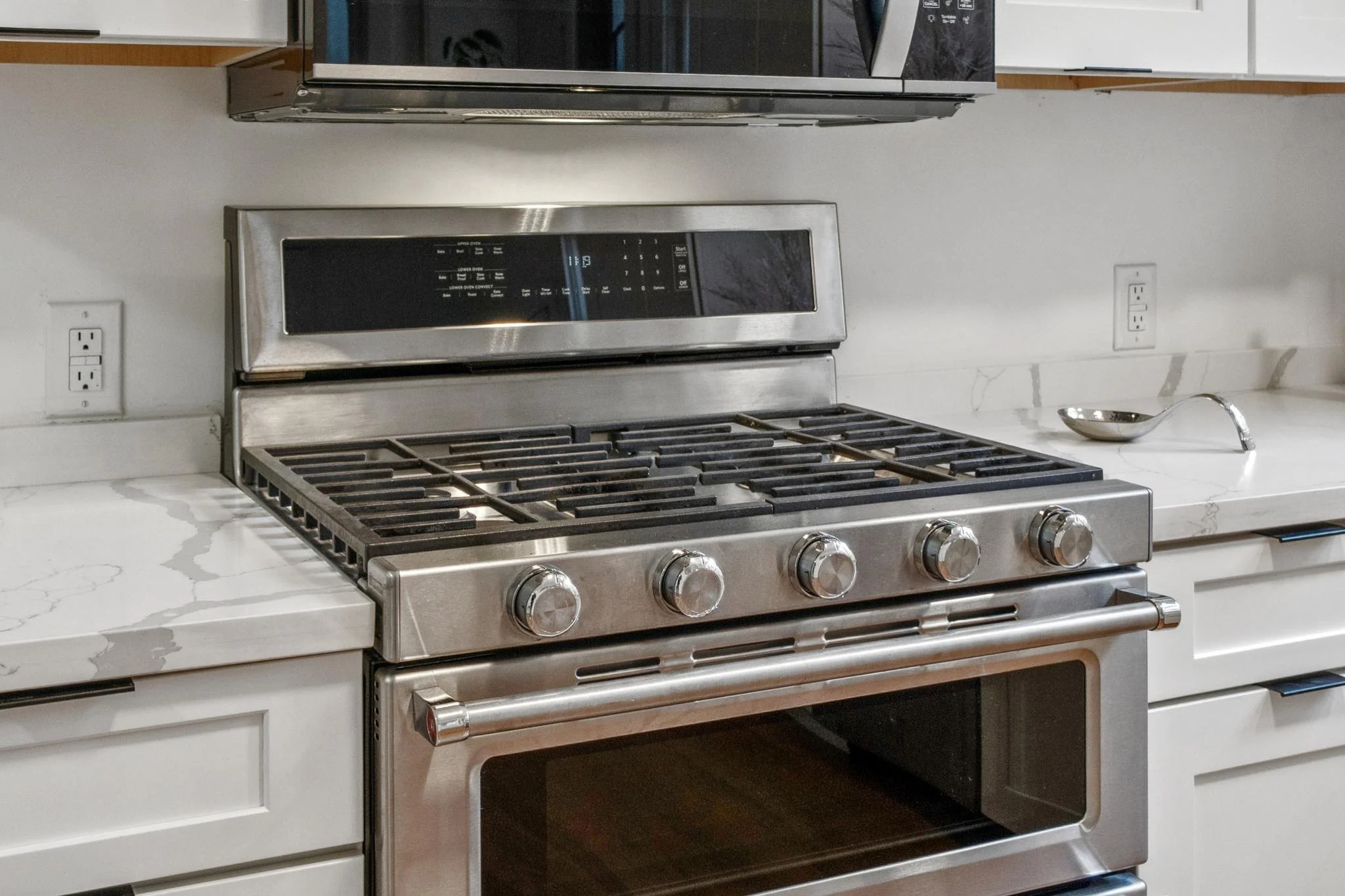 Stainless steel gas stove with five burners, black control knobs, and an oven door at the bottom. White marble countertops on either side and a microwave above. Two electrical outlets on the wall, one to the left and one to the right.