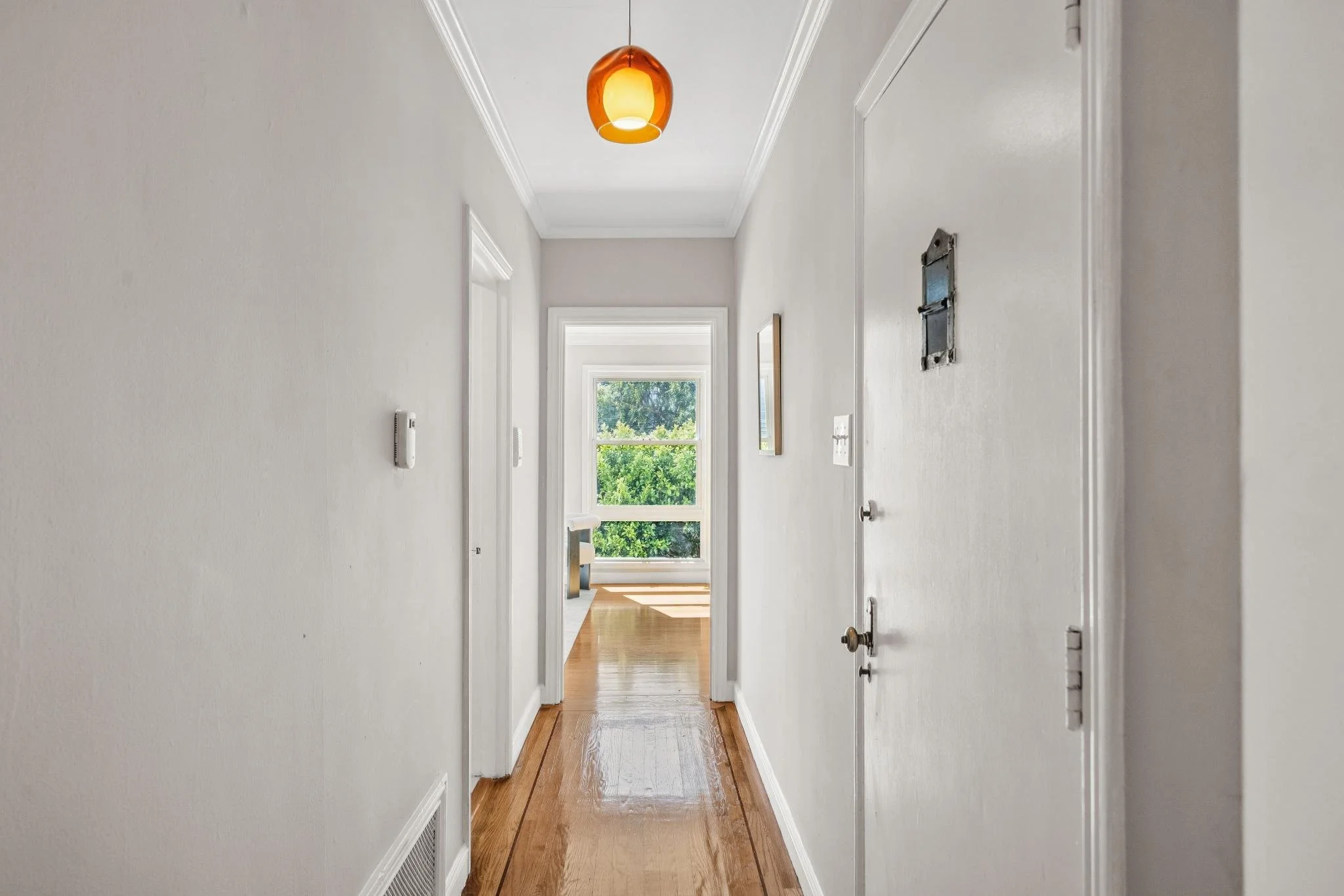 A bright, narrow hallway with white walls and wooden flooring, lit by an orange pendant lamp, leading to a room with large windows overlooking green trees.