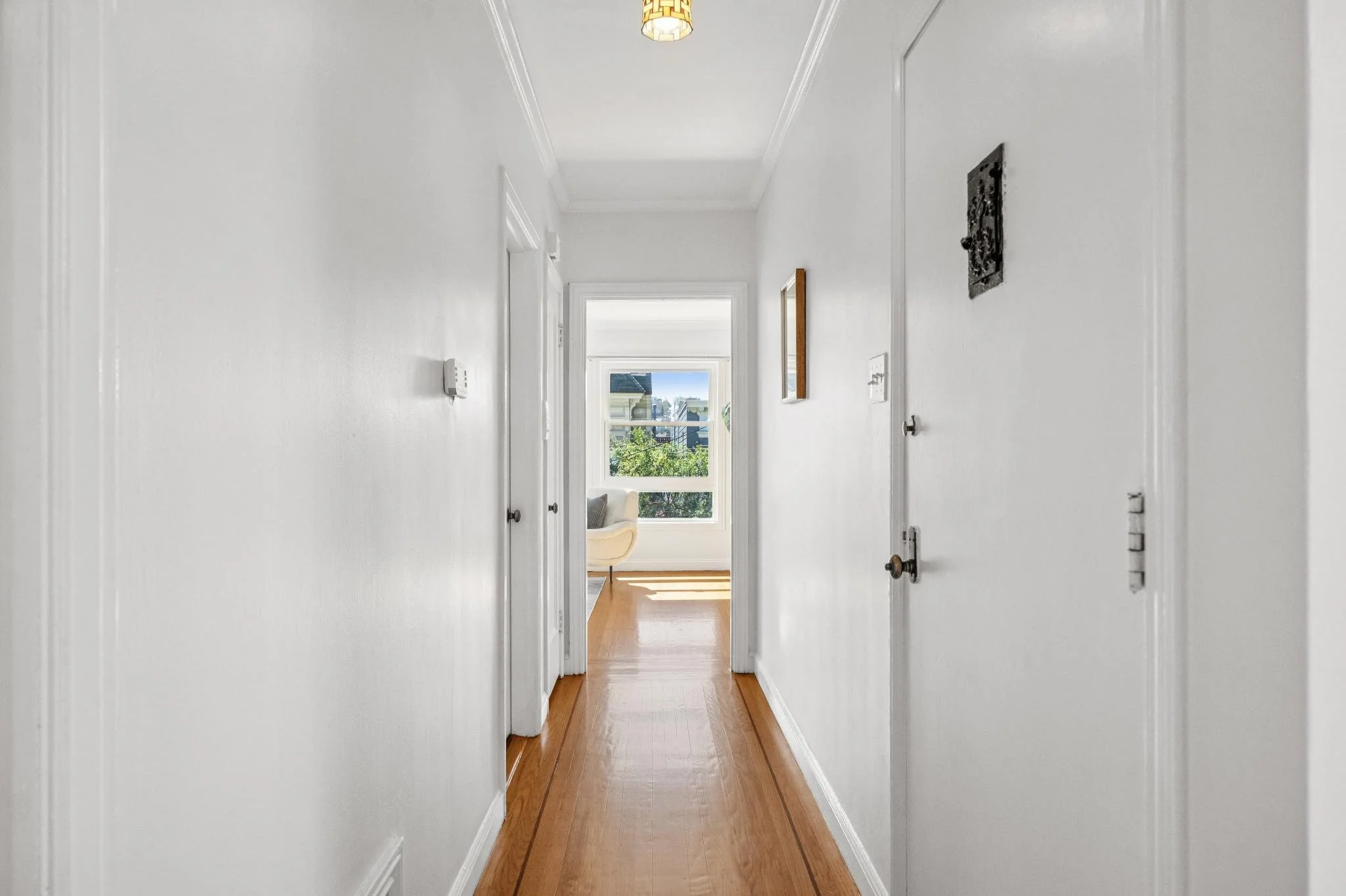 Bright hallway with white walls, wooden floor, and closed doors on either side, leading to a sunny room with a large window and a armchair.