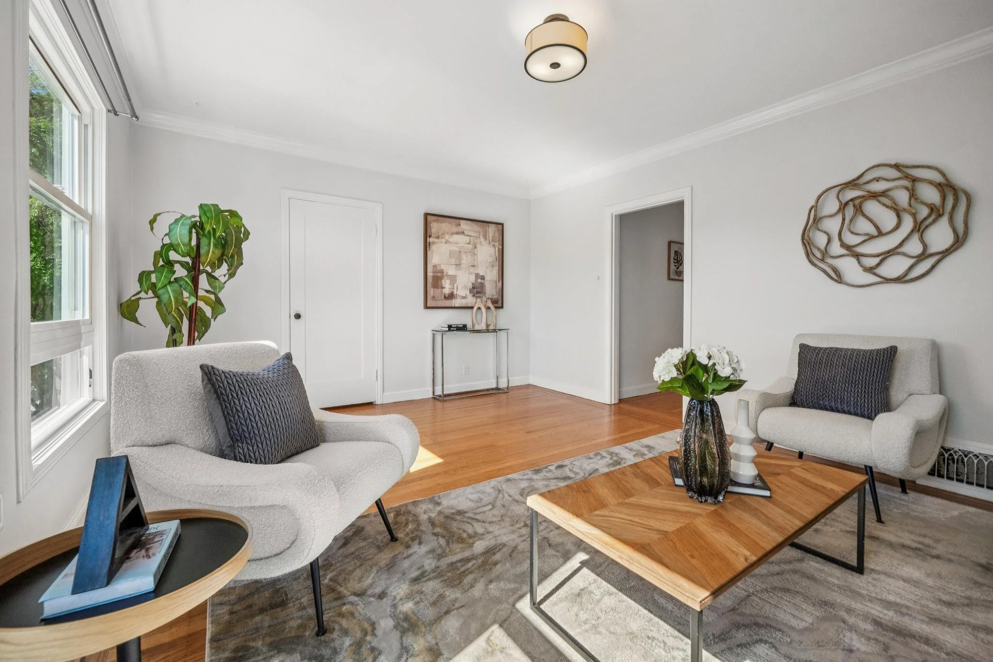 Living room with white walls, hardwood floors, two white armchairs with black pillows, a wooden coffee table with a vase of white flowers, a large potted plant near the window, wall art, and decorative objects on a console table.