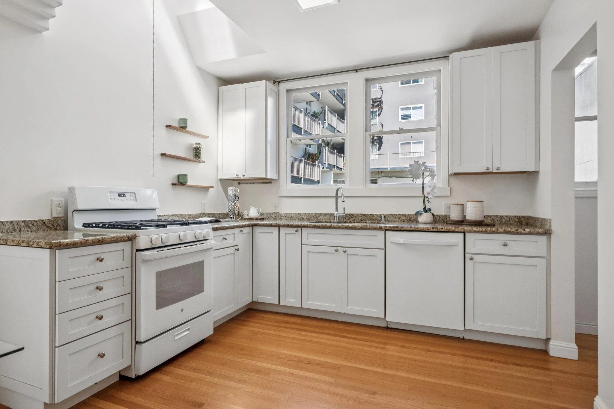 White kitchen with wood flooring, granite countertops, white cabinets, stove, sink, and large window overlooking apartment building balconies.