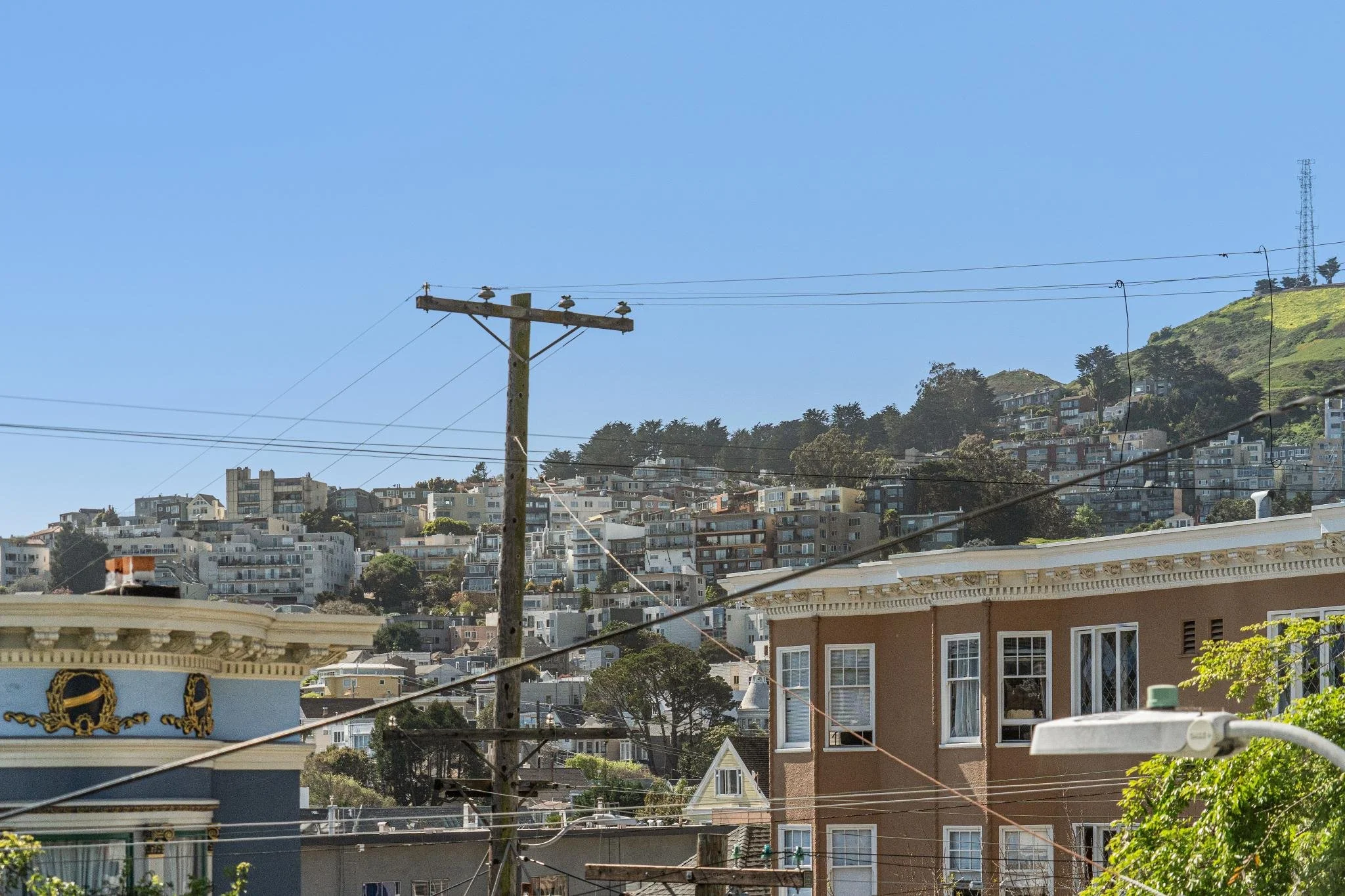 Cityscape with residential buildings on a hillside, telephone pole, several power lines, and a streetlamp in foreground, with a clear blue sky.
